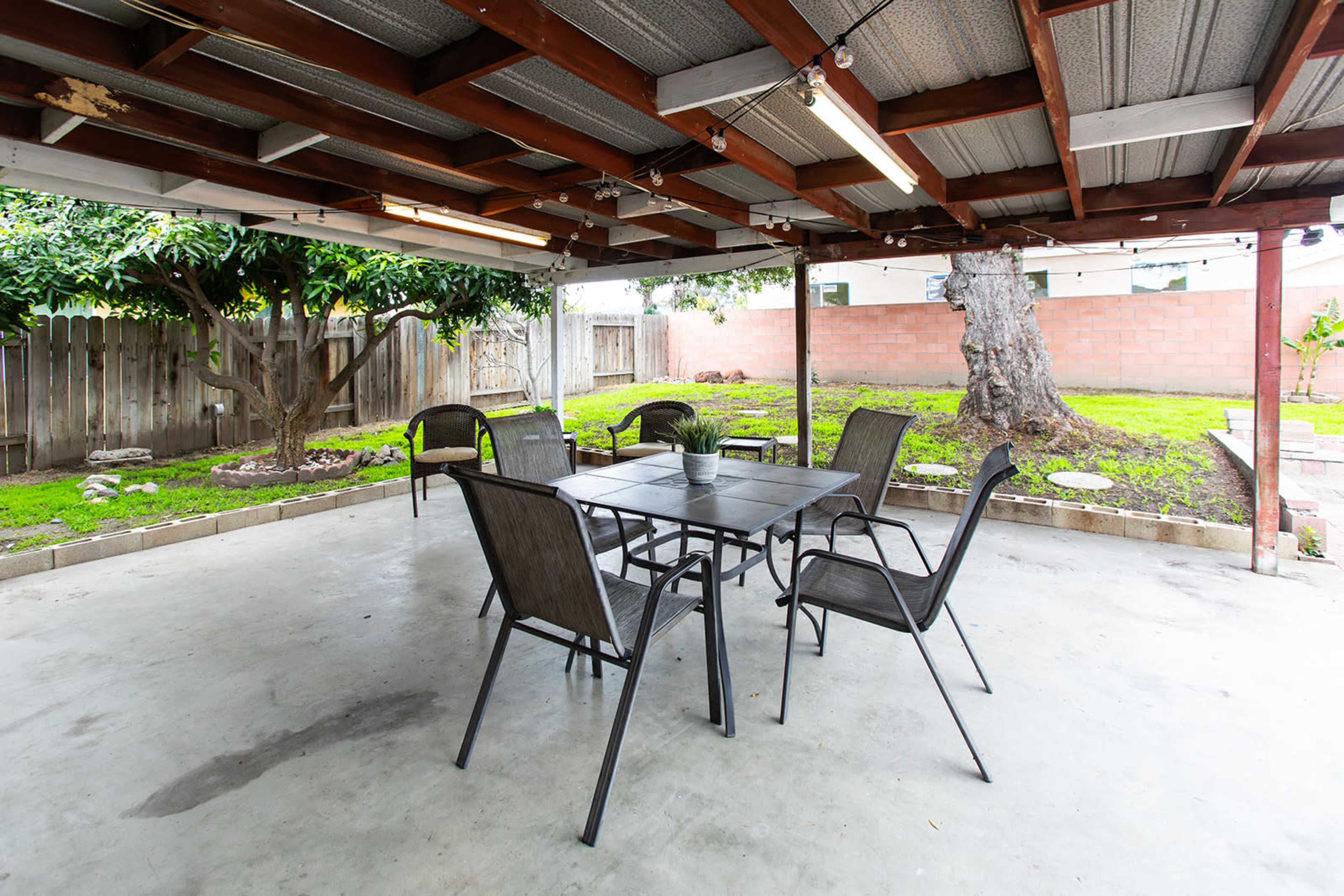 A covered outdoor patio with a table and several chairs, surrounded by a backyard with grass and trees.