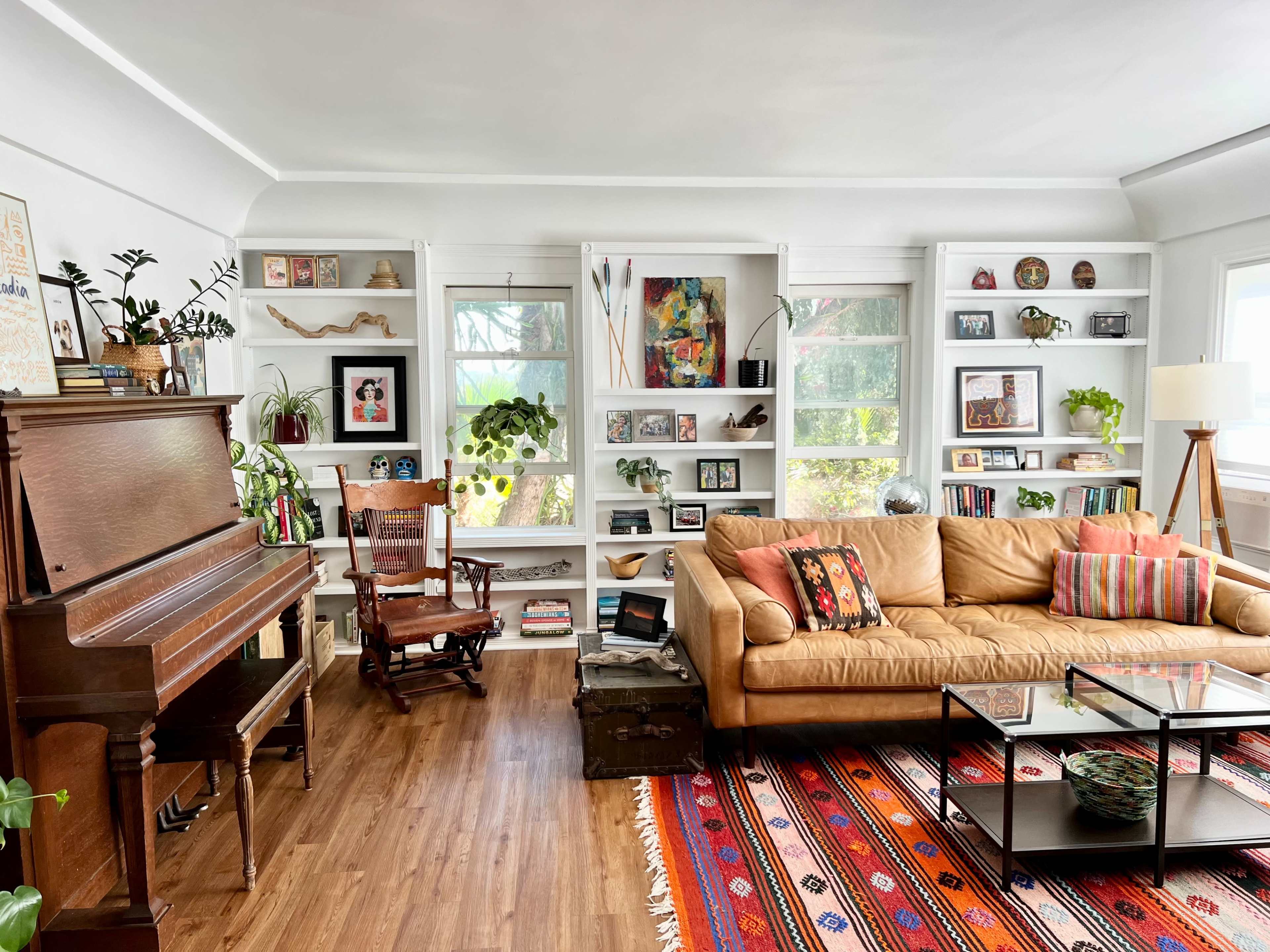 A cozy living room featuring a piano, an armchair, a beige couch with decorative pillows, and shelves filled with various decorations and plants.