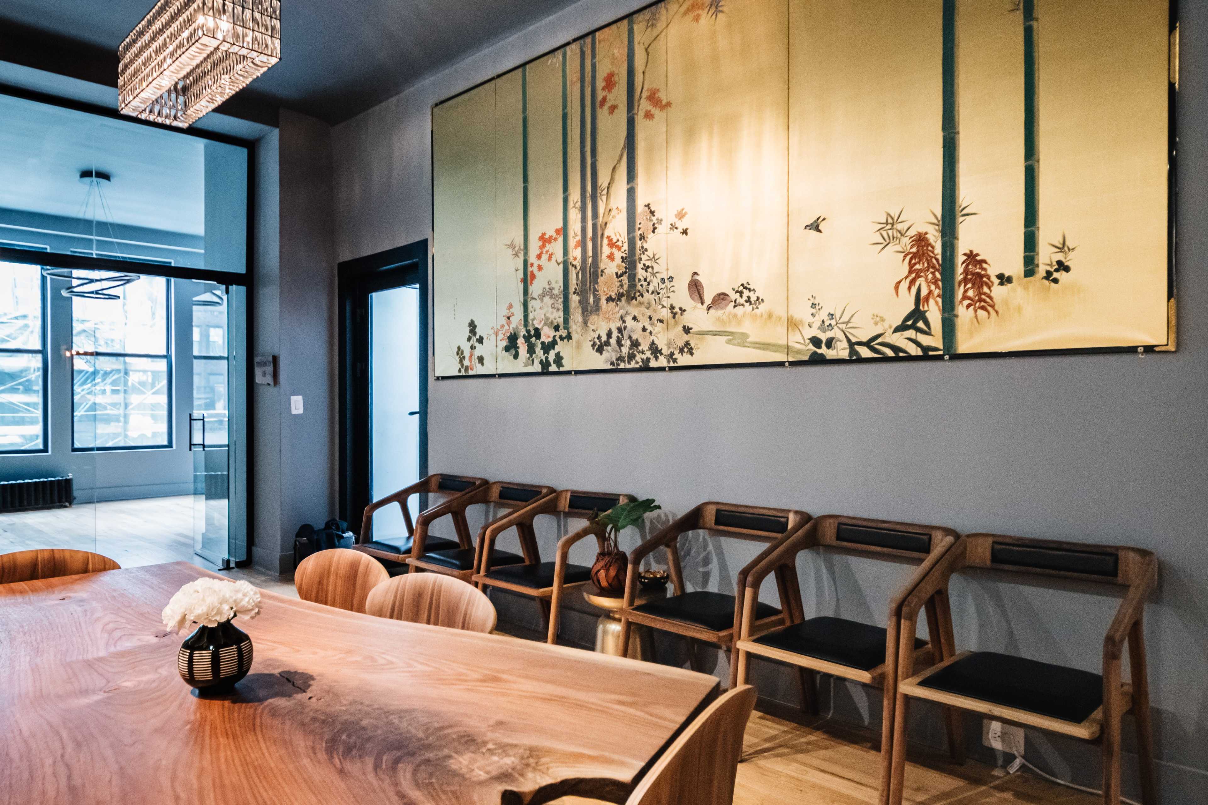 A wooden dining table is surrounded by chairs in a room featuring a large decorative wall hanging of bamboo and flowers.