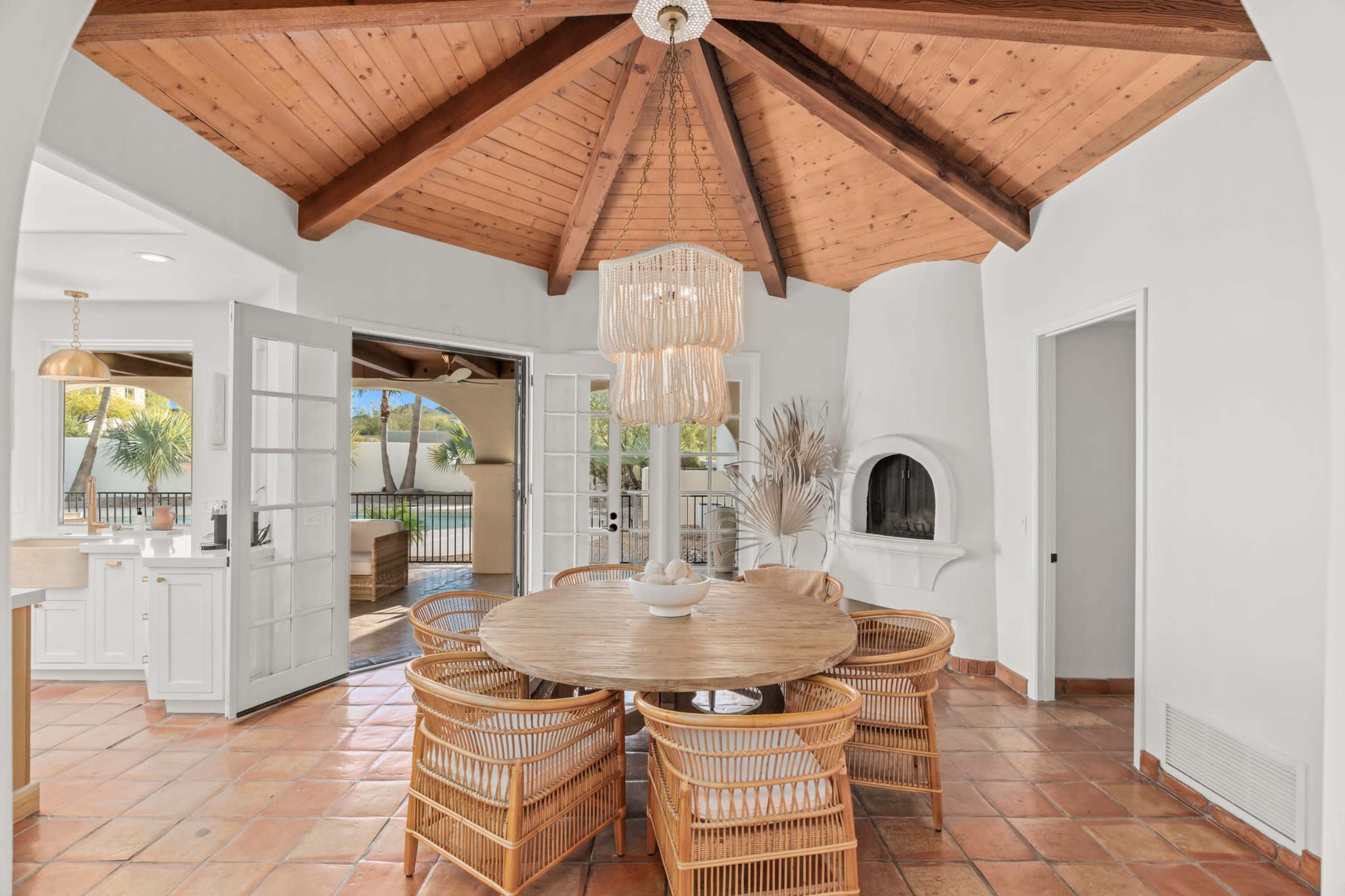 A dining area featuring a round wooden table surrounded by wicker chairs, with a vaulted wooden ceiling and large open doors leading to an outdoor space.