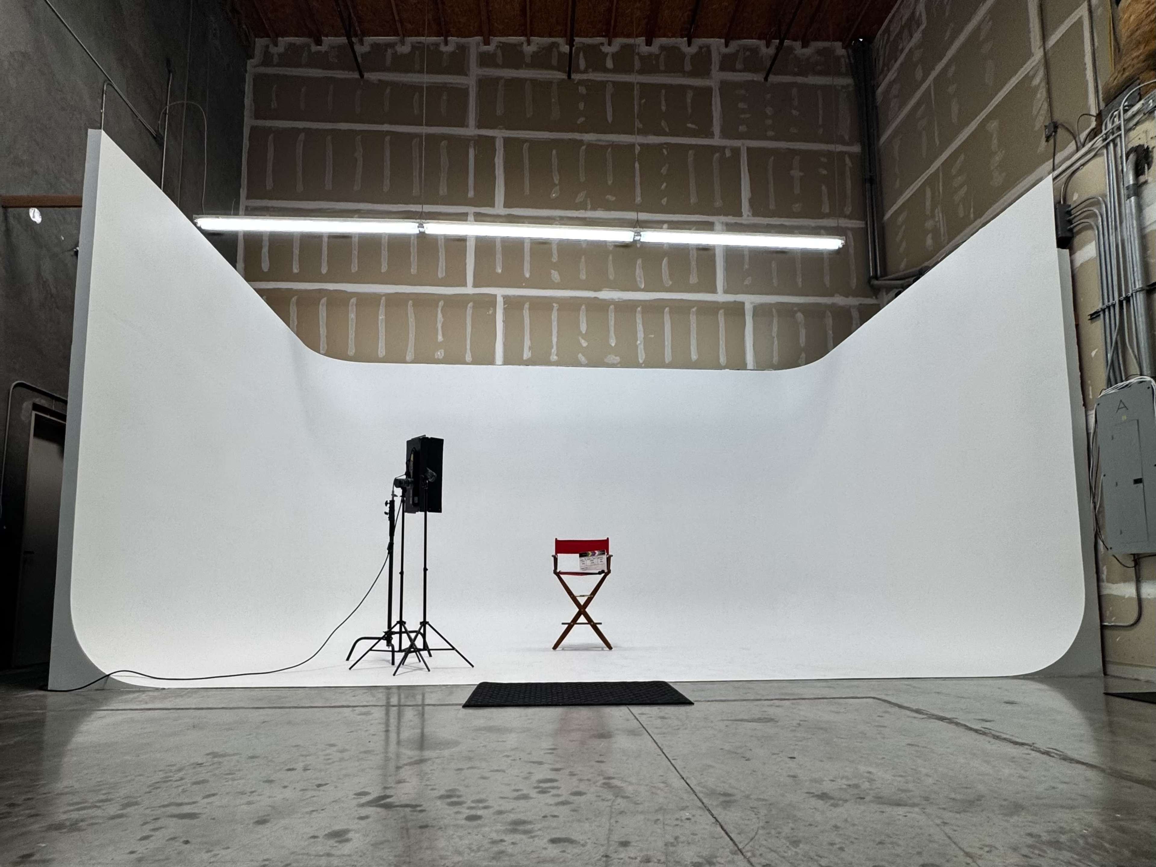 A photography studio features a large white backdrop, a light stand with a softbox, and a red director's chair positioned on a gray floor.
