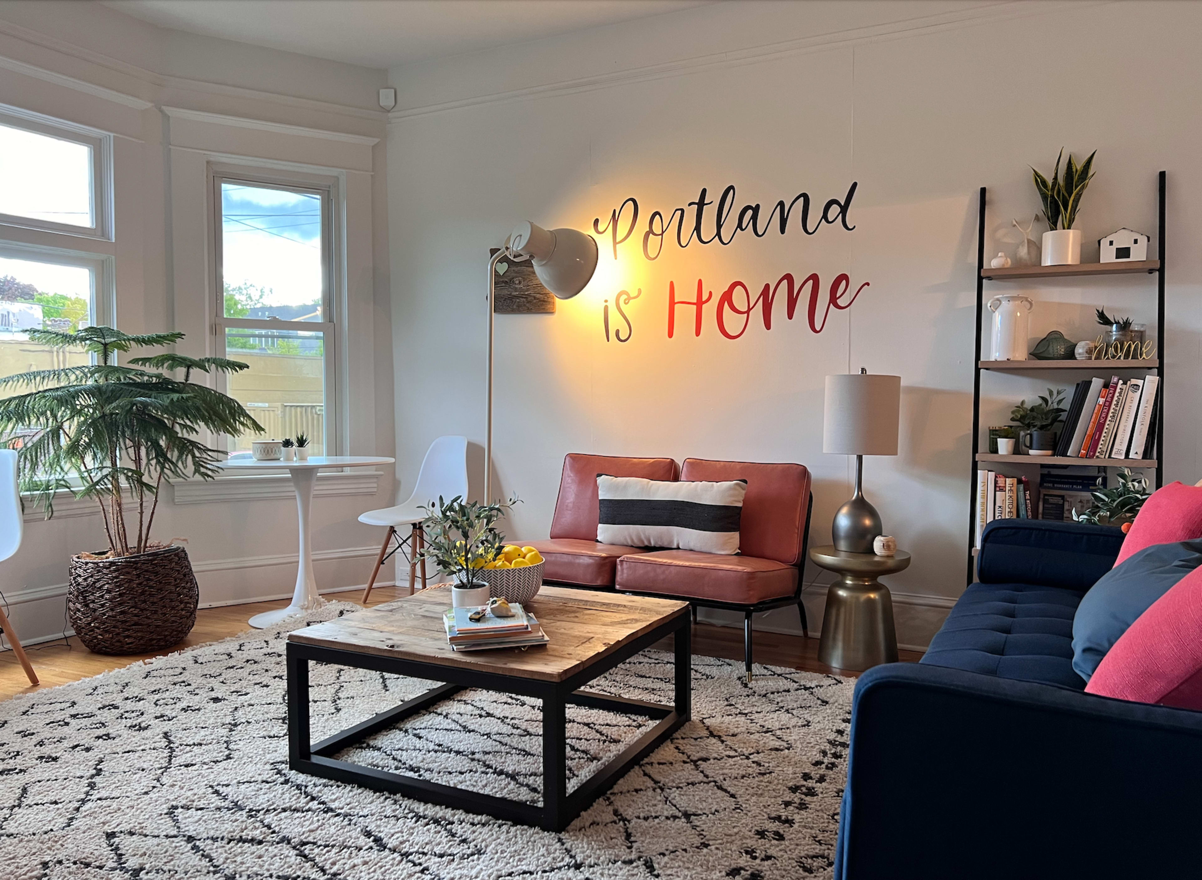 The image shows a stylish living room with a large coffee table, a pink couch, a blue couch, and a wall decorated with the phrase "Portland is Home."