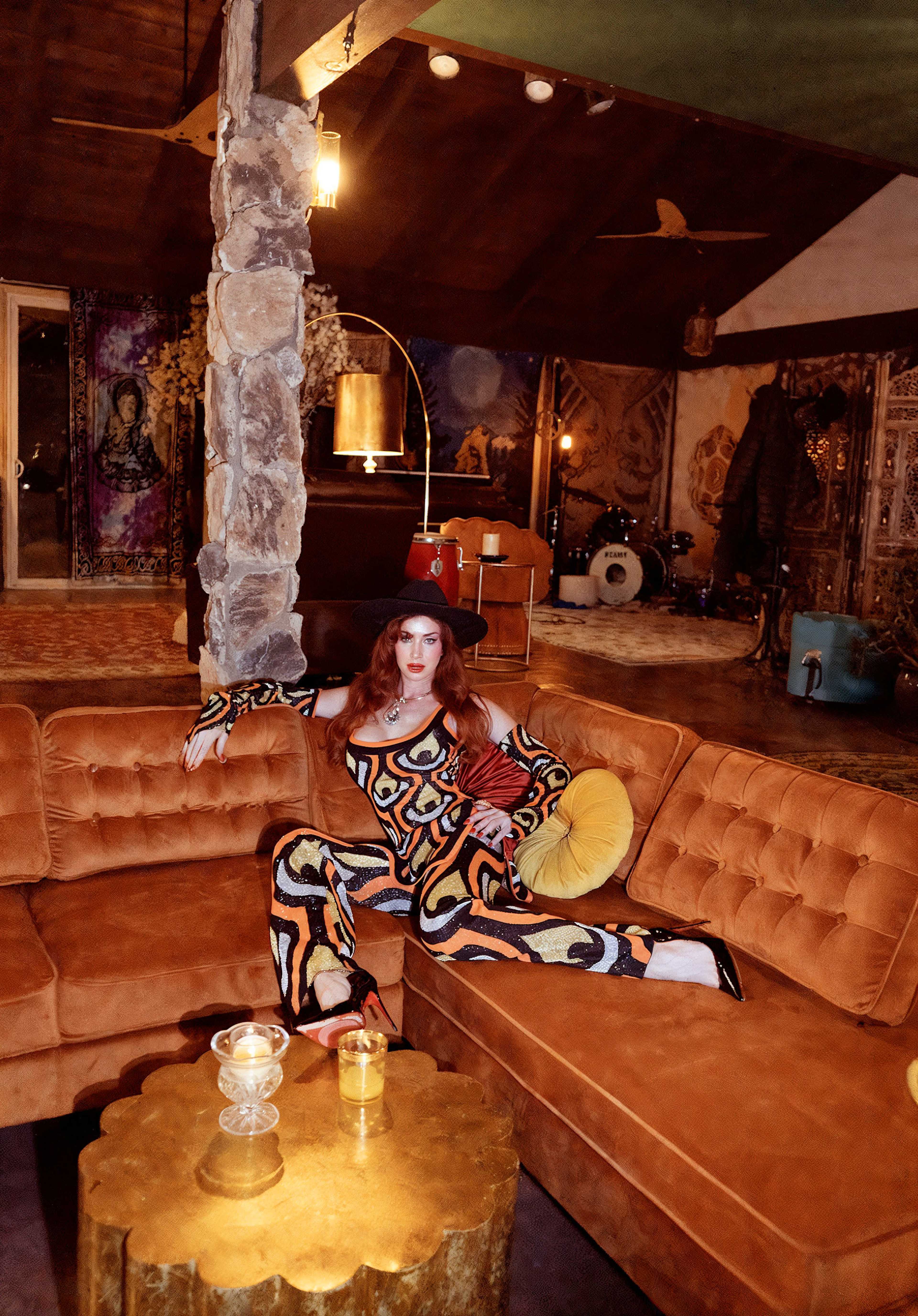 A woman in a colorful patterned outfit sits on an orange couch in a stylishly decorated room with artistic elements and a drum set in the background.
