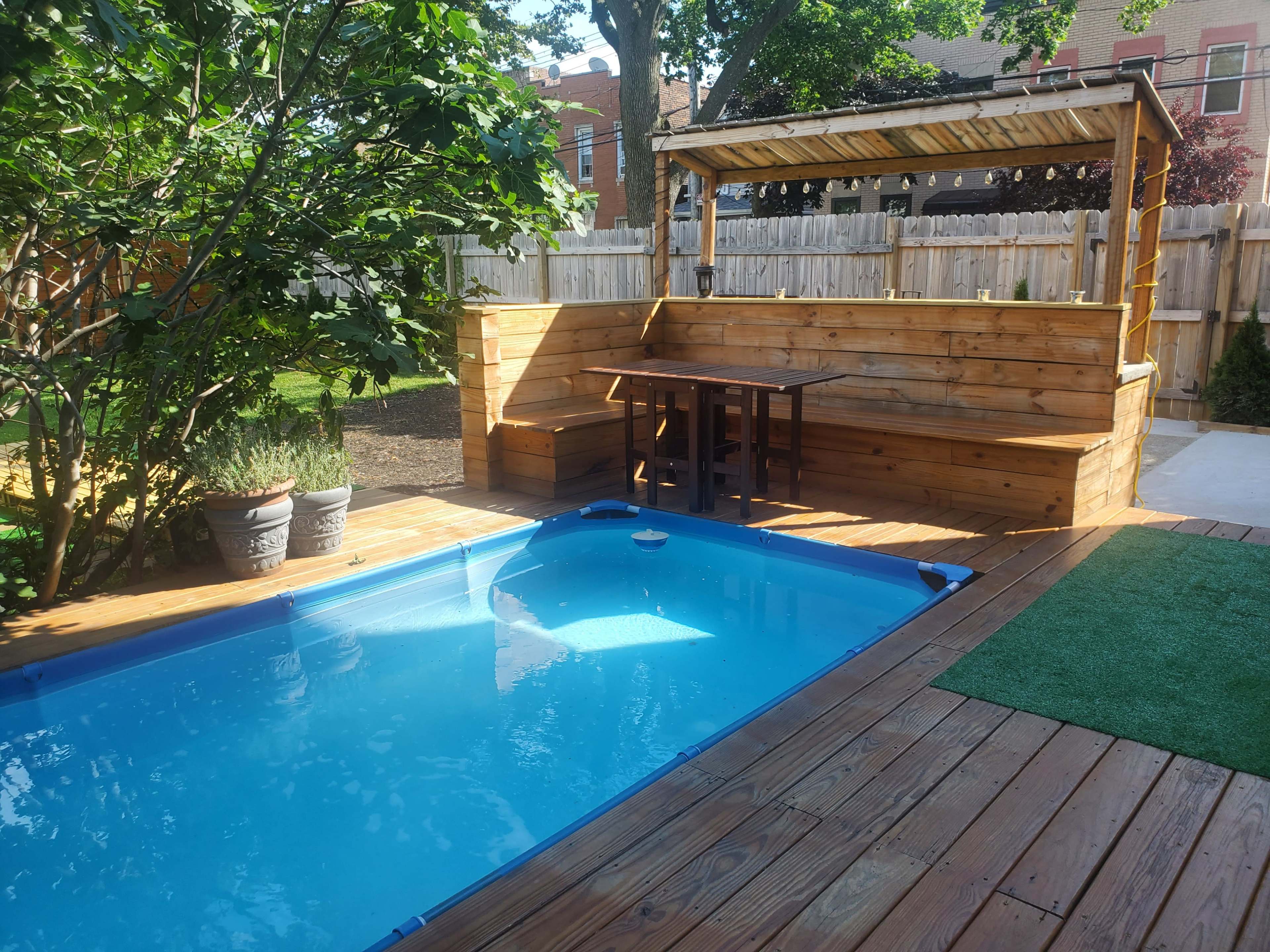 A small backyard featuring a blue pool, a wooden seating area with a table, and green plants within a fenced enclosure.