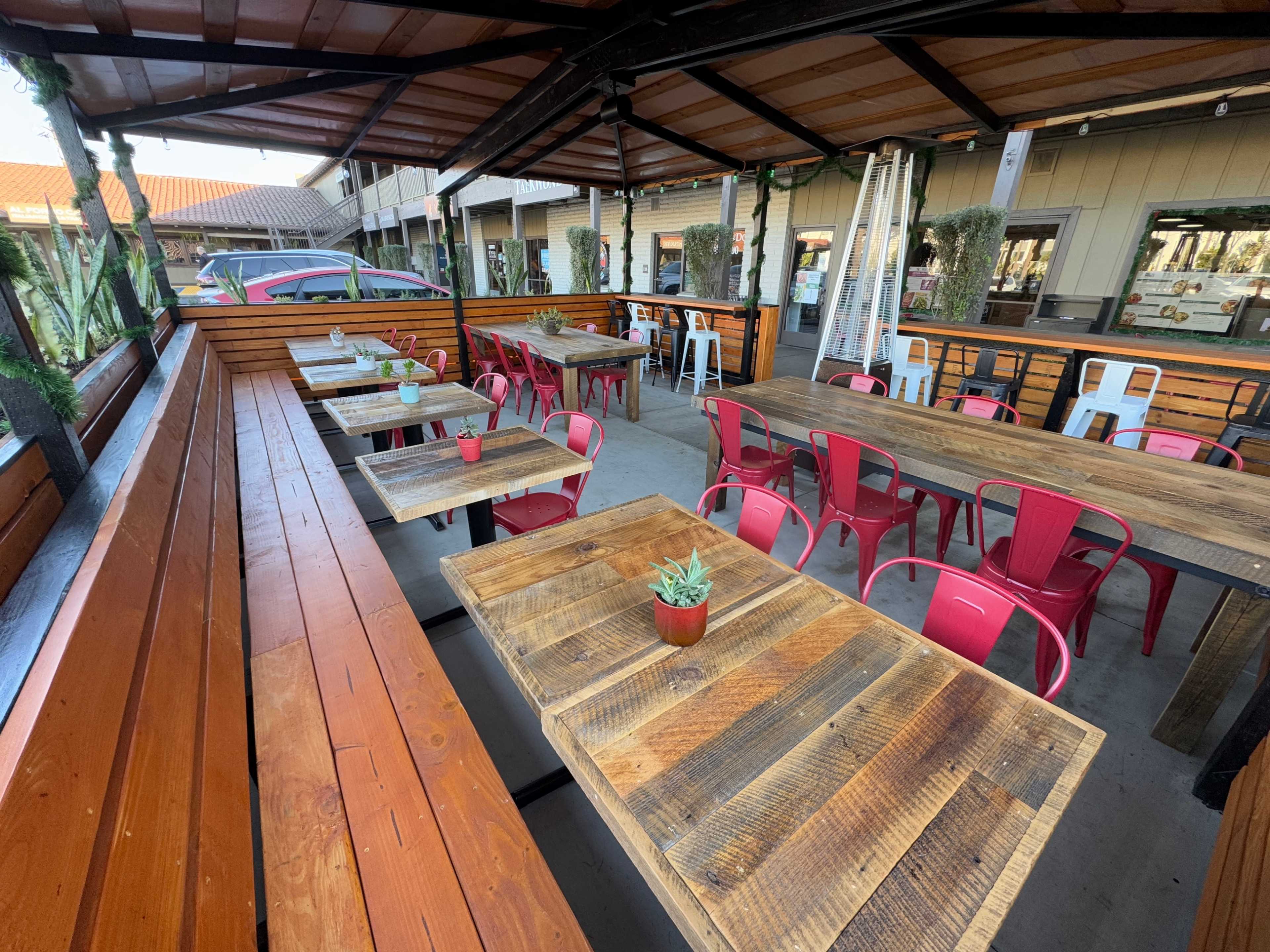 The image shows an outdoor dining area with wooden tables, metal pink chairs, and a canopy covering.