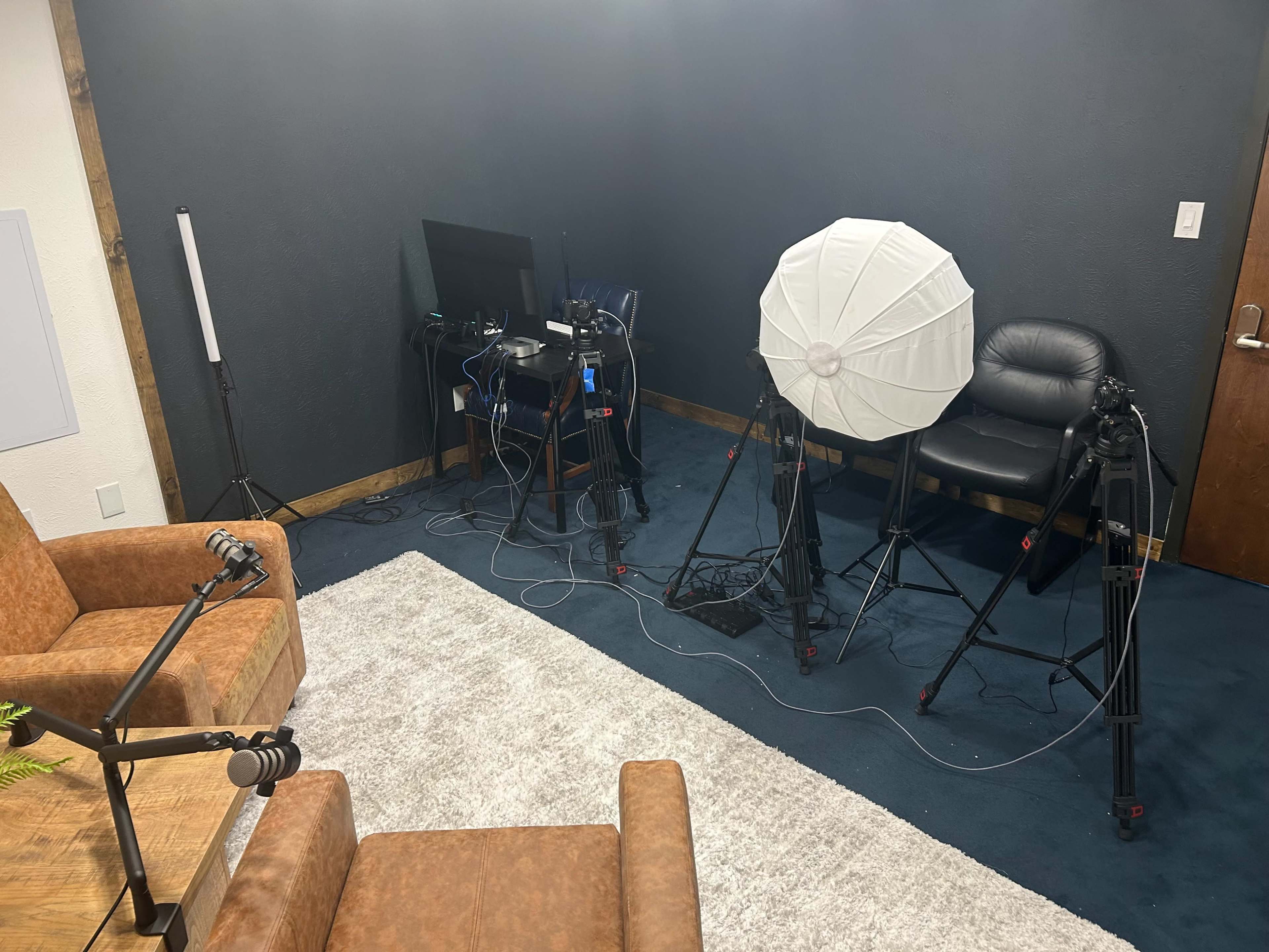 The image shows a recording studio setup with two brown chairs, a light umbrella, several camera tripods, and a computer monitor.