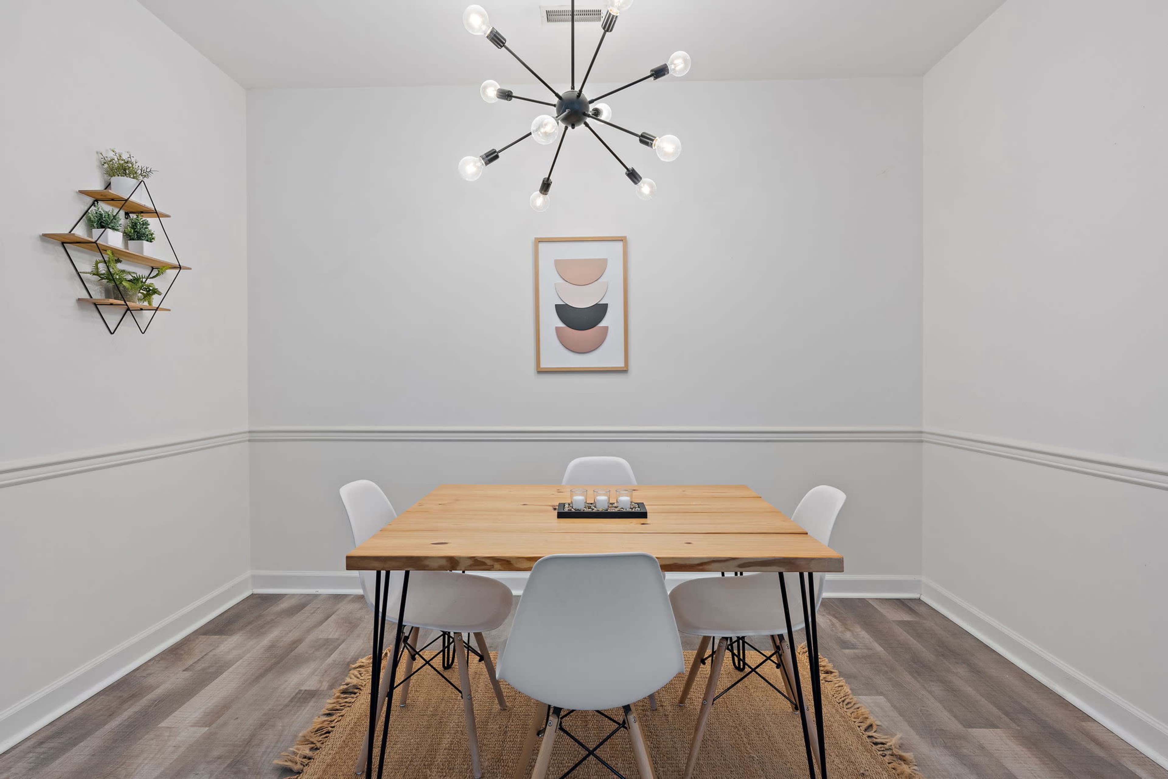 A minimalist dining room features a wooden table surrounded by white chairs, with a modern light fixture overhead and a framed artwork on the wall.
