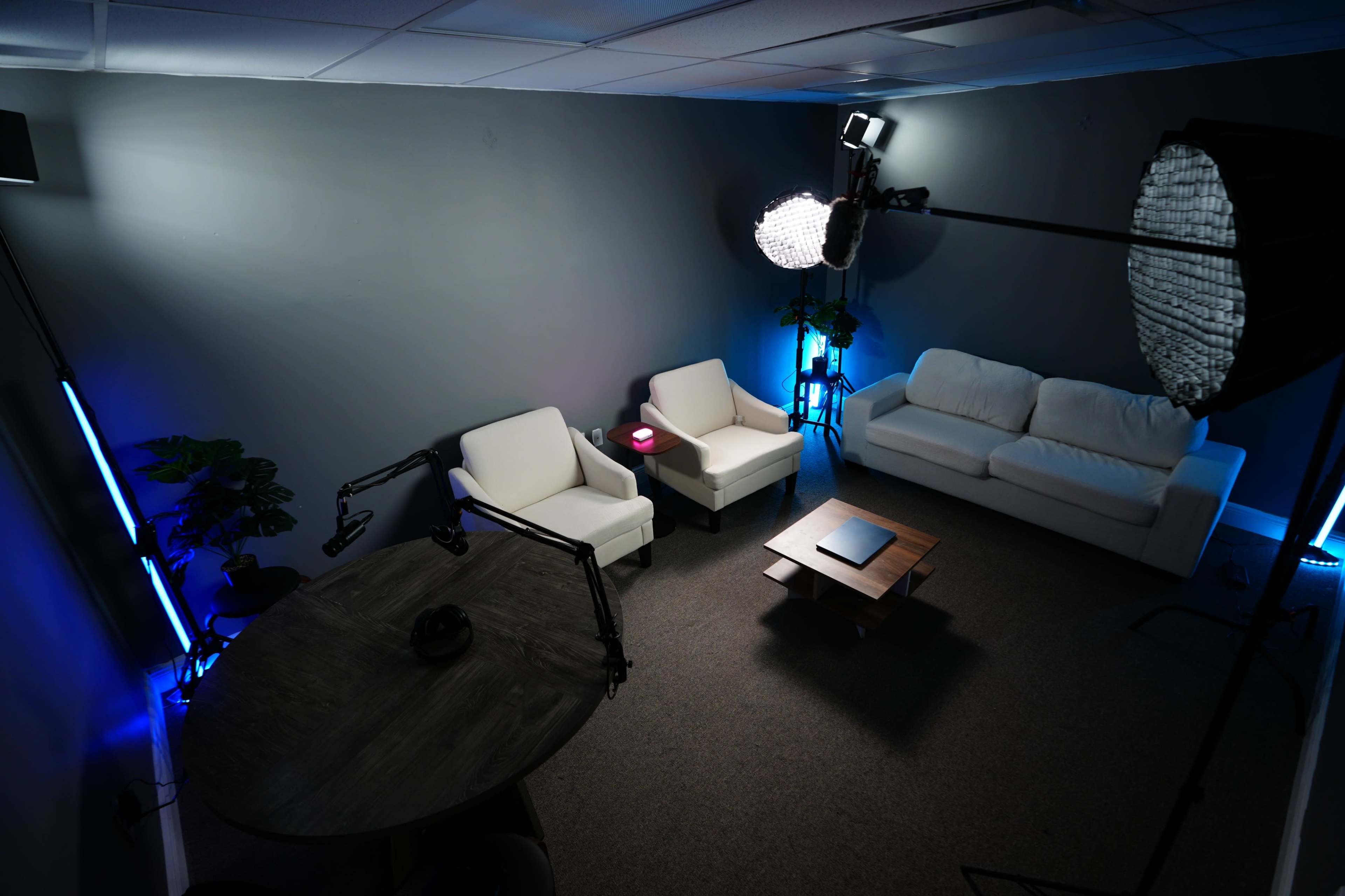 The image shows a corner of a modern, minimalistic lounge area with two white armchairs and a white sofa arranged around a coffee table, illuminated by blue and soft white lighting.