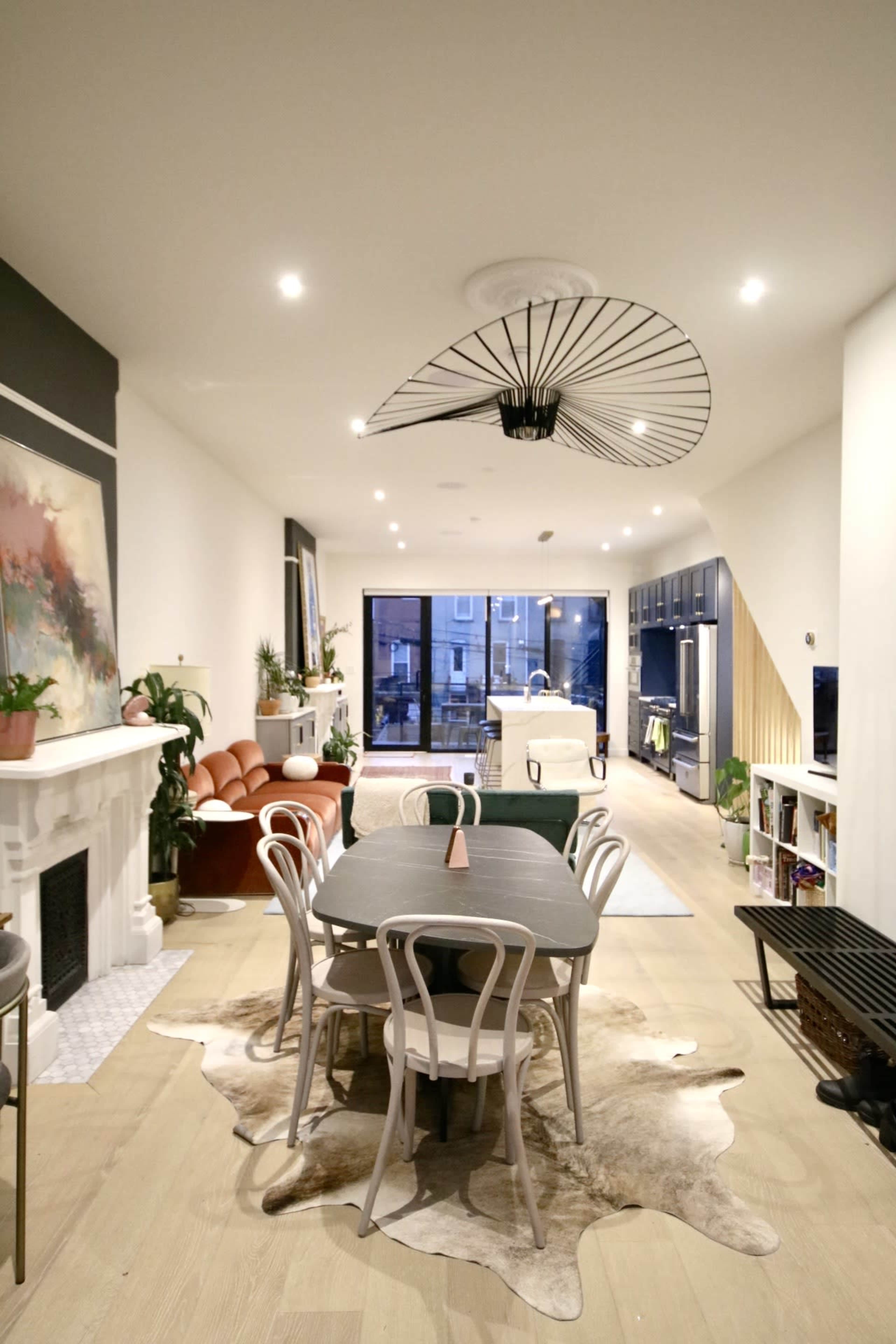 The image shows a modern living and dining area featuring a large dining table with chairs, a fireplace, potted plants, and a glass door leading to an outdoor space.
