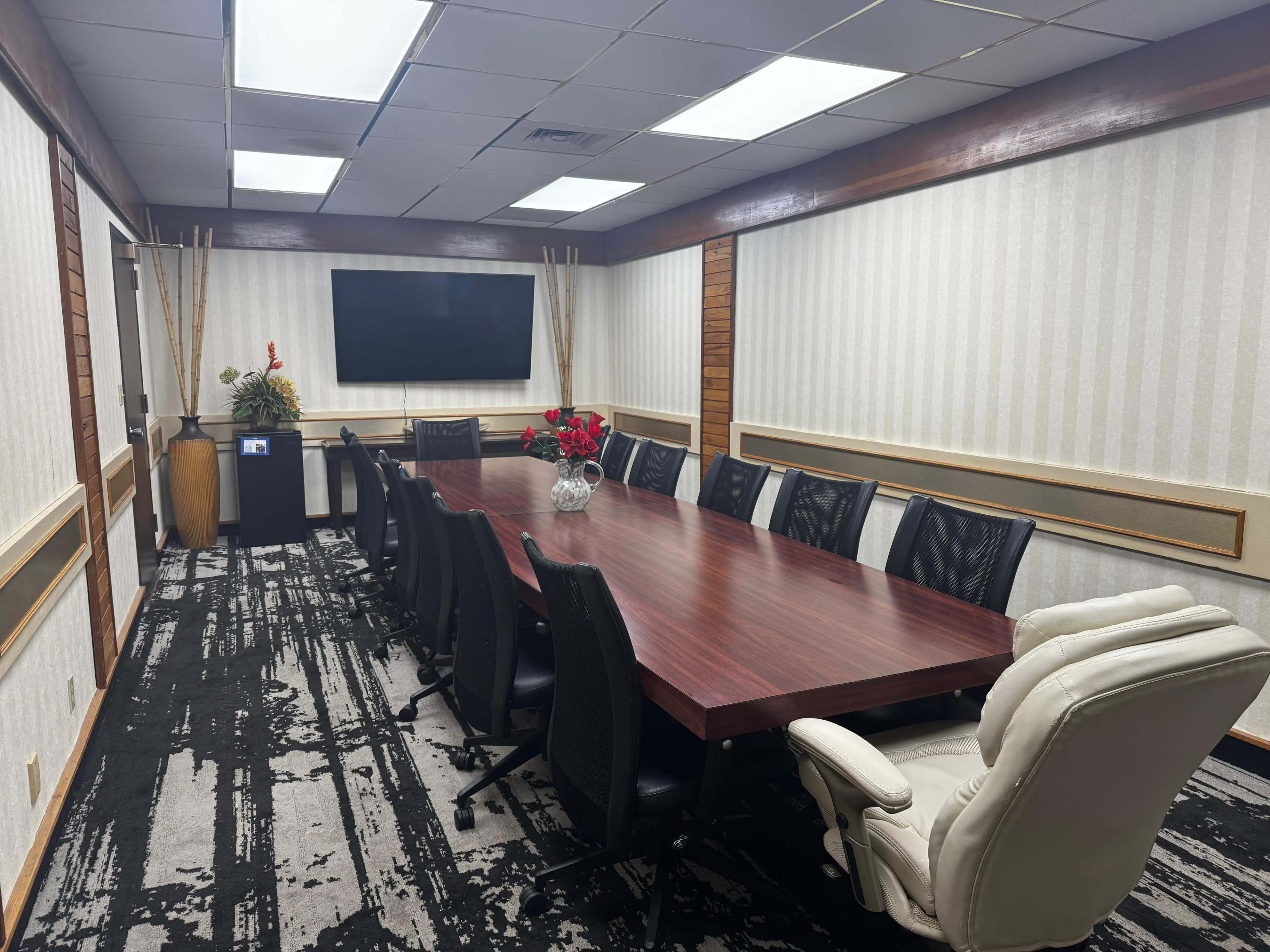 The image depicts a conference room with a large wooden table surrounded by black and white chairs, featuring a wall-mounted television and decorative plants.
