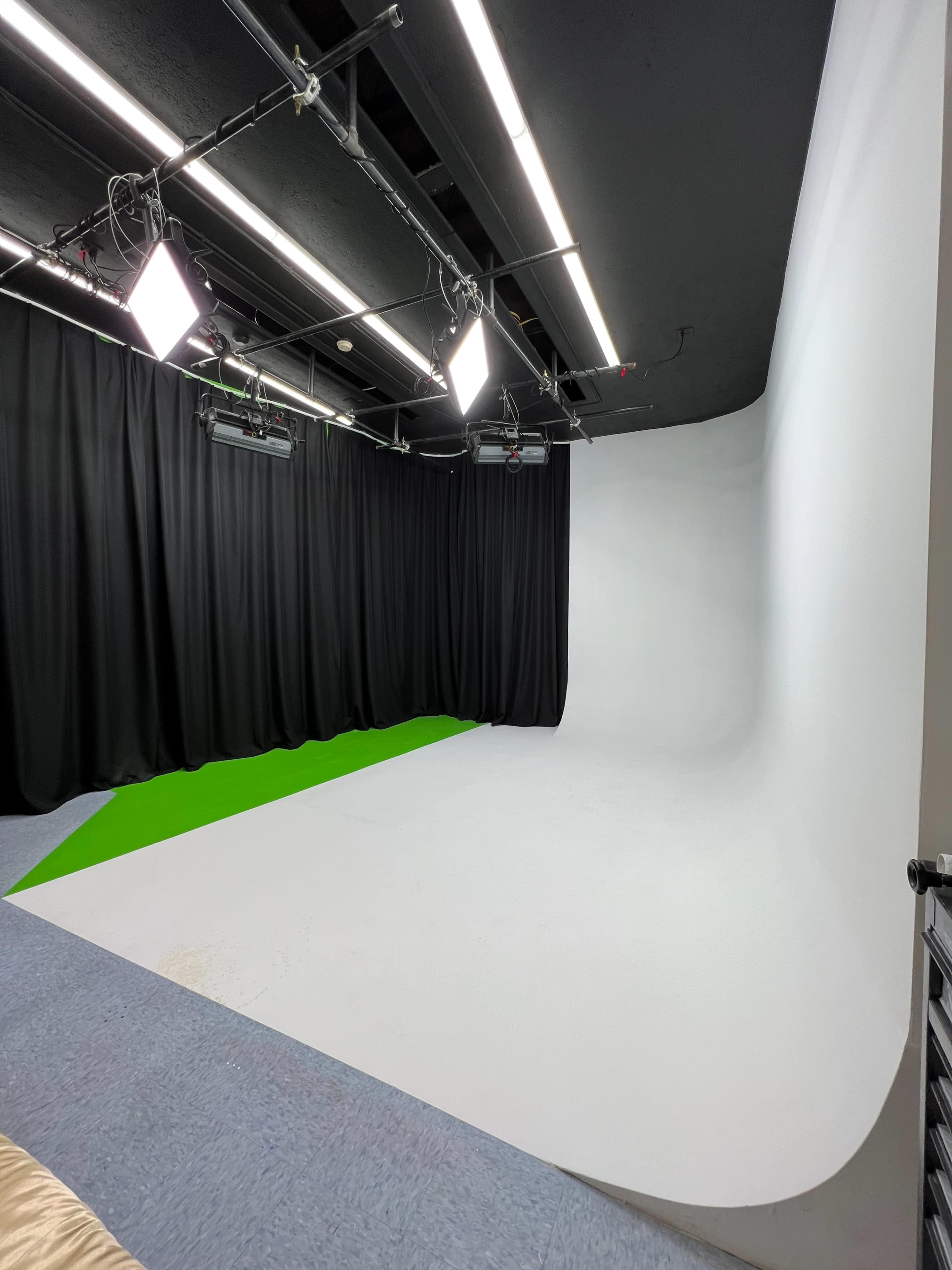 The image shows a spacious studio with a curved white backdrop, green flooring, and softbox lights, surrounded by black curtains.
