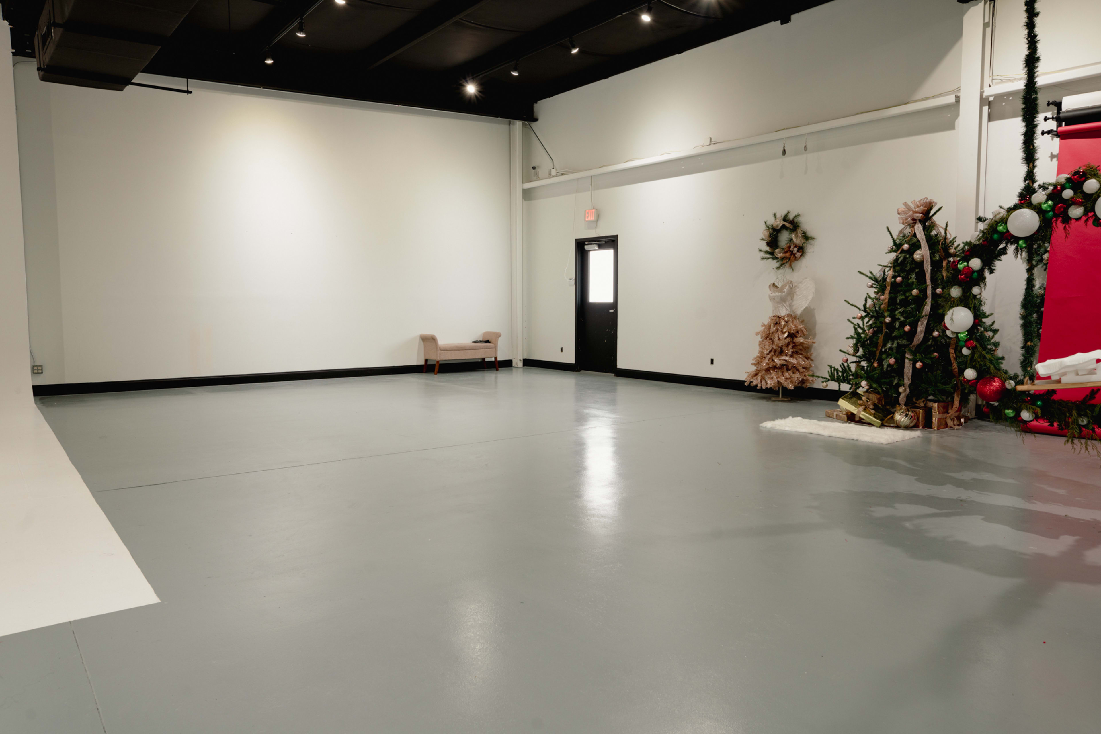 The image shows a spacious, empty room with a light gray floor, a single chair against one wall, and holiday decorations in one corner.