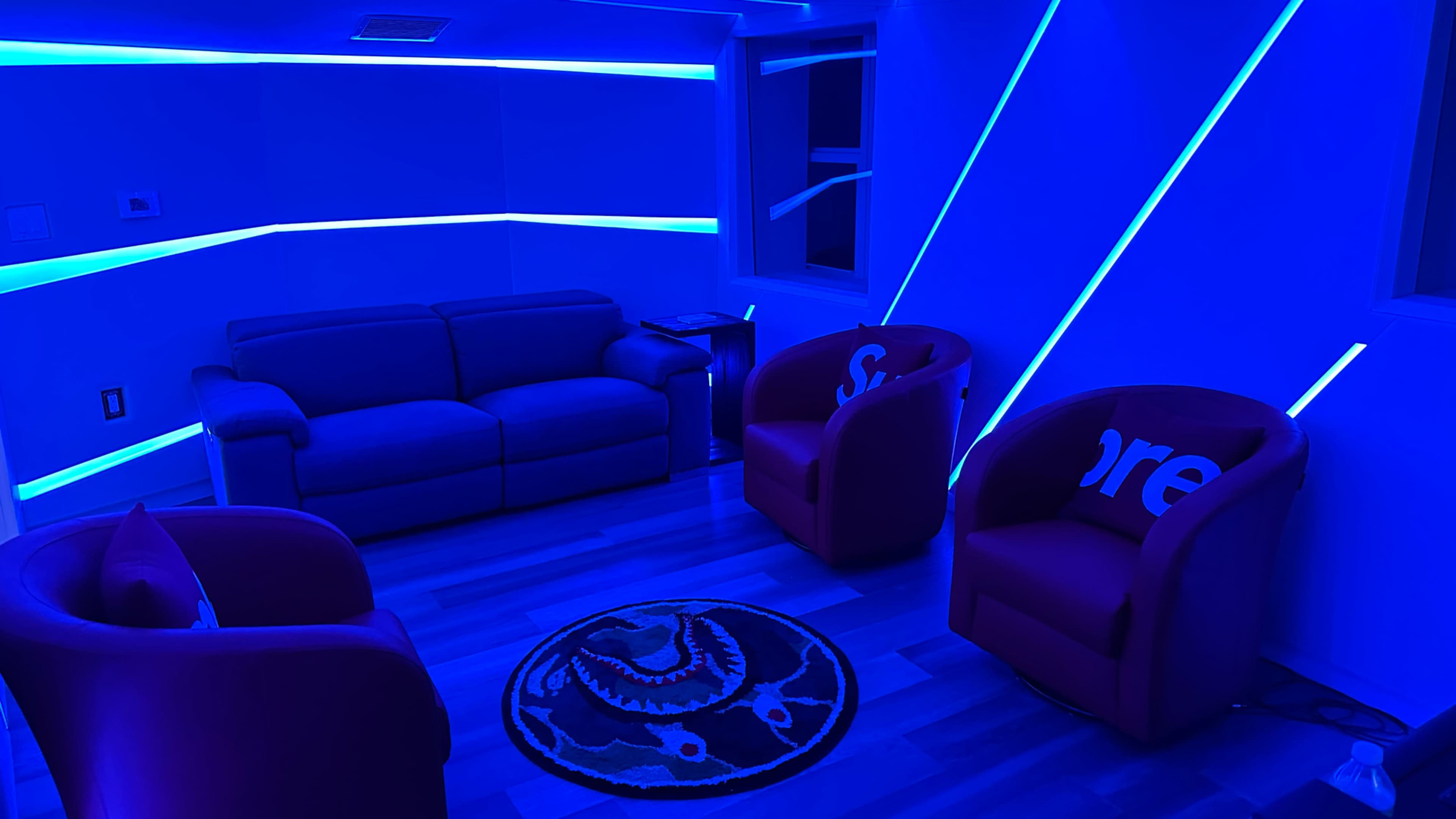 A modern room features a blue-lit sofa and two red chairs with decorative throw pillows, arranged around a circular rug.