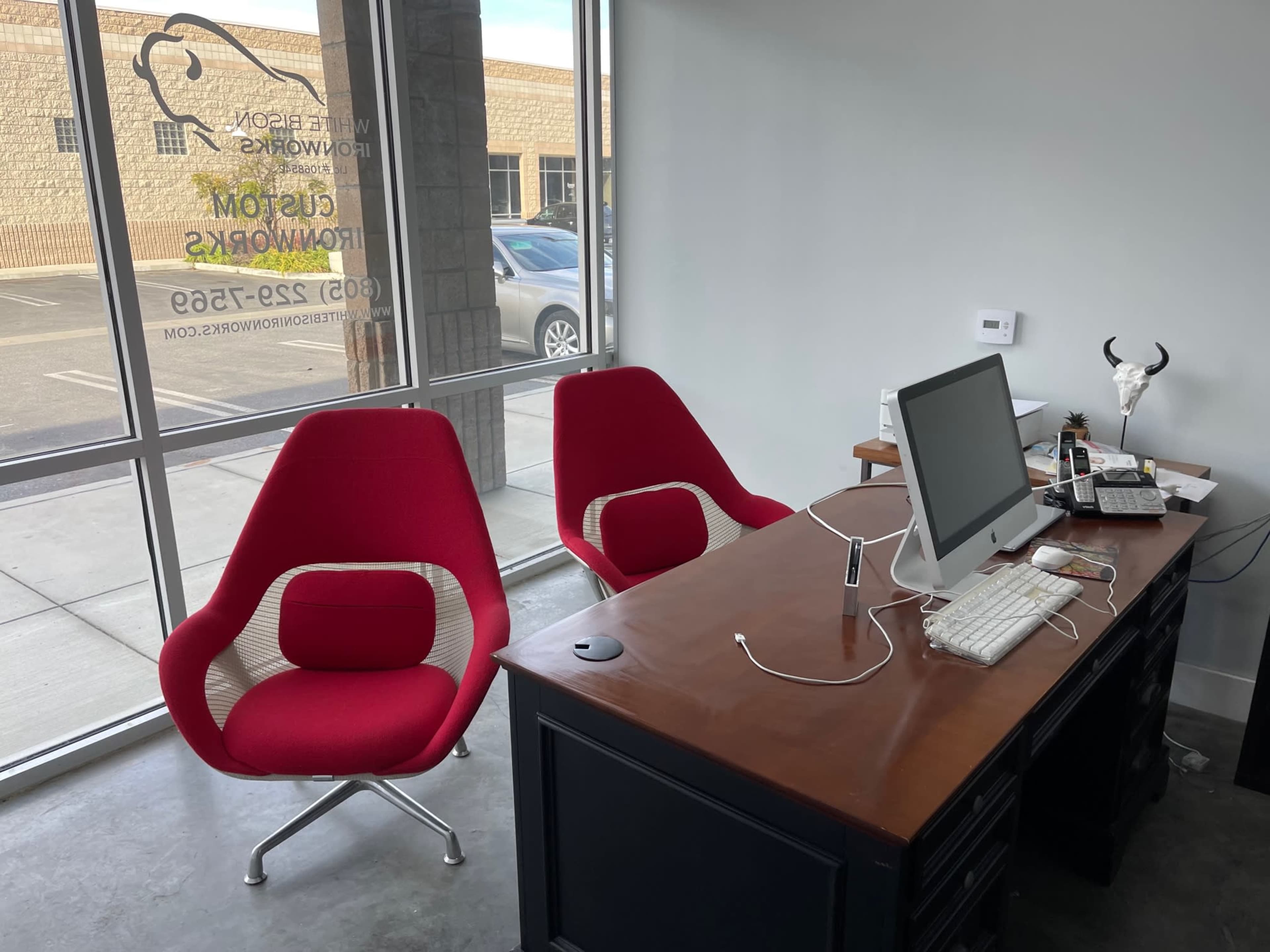 A modern office space with two red chairs facing a wooden desk that holds a computer and various office supplies, set against a large window.