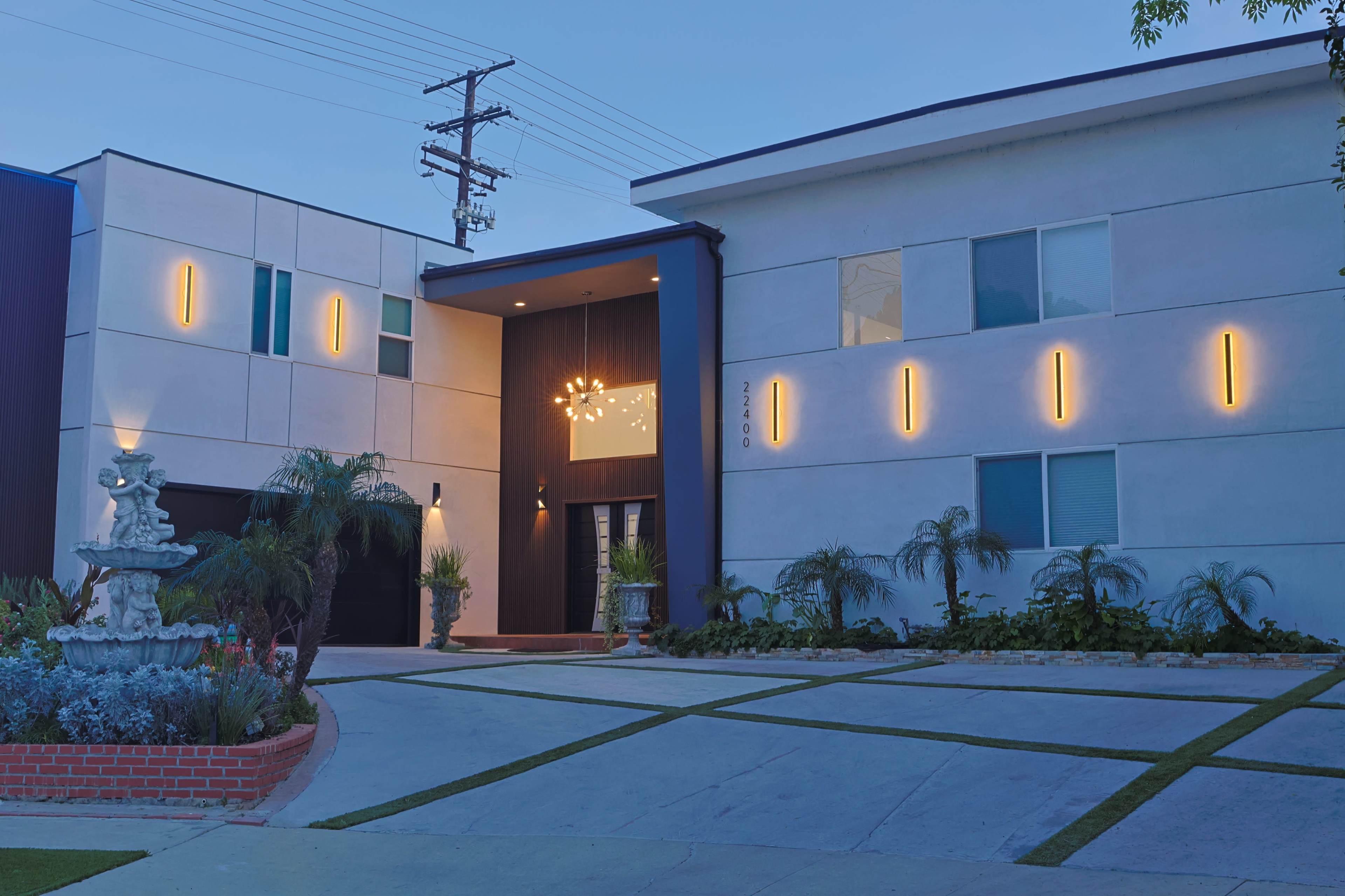 The image shows a modern two-story house with illuminated vertical lights along its exterior, a landscaped front yard, and a driveway leading to a garage.