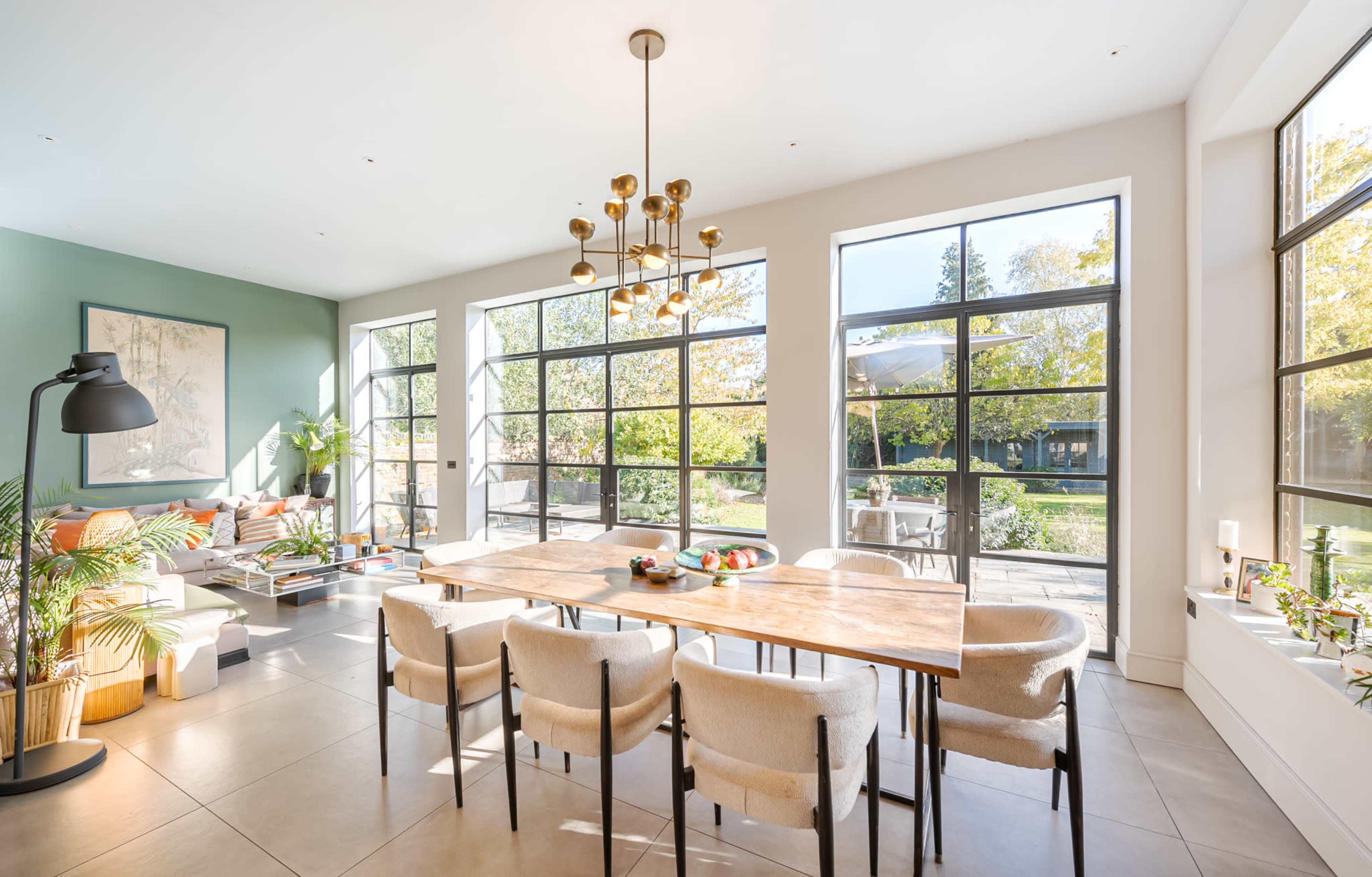 The image shows a modern, well-lit dining area with large windows, a wooden dining table surrounded by upholstered chairs, and a view of a green outdoor space.