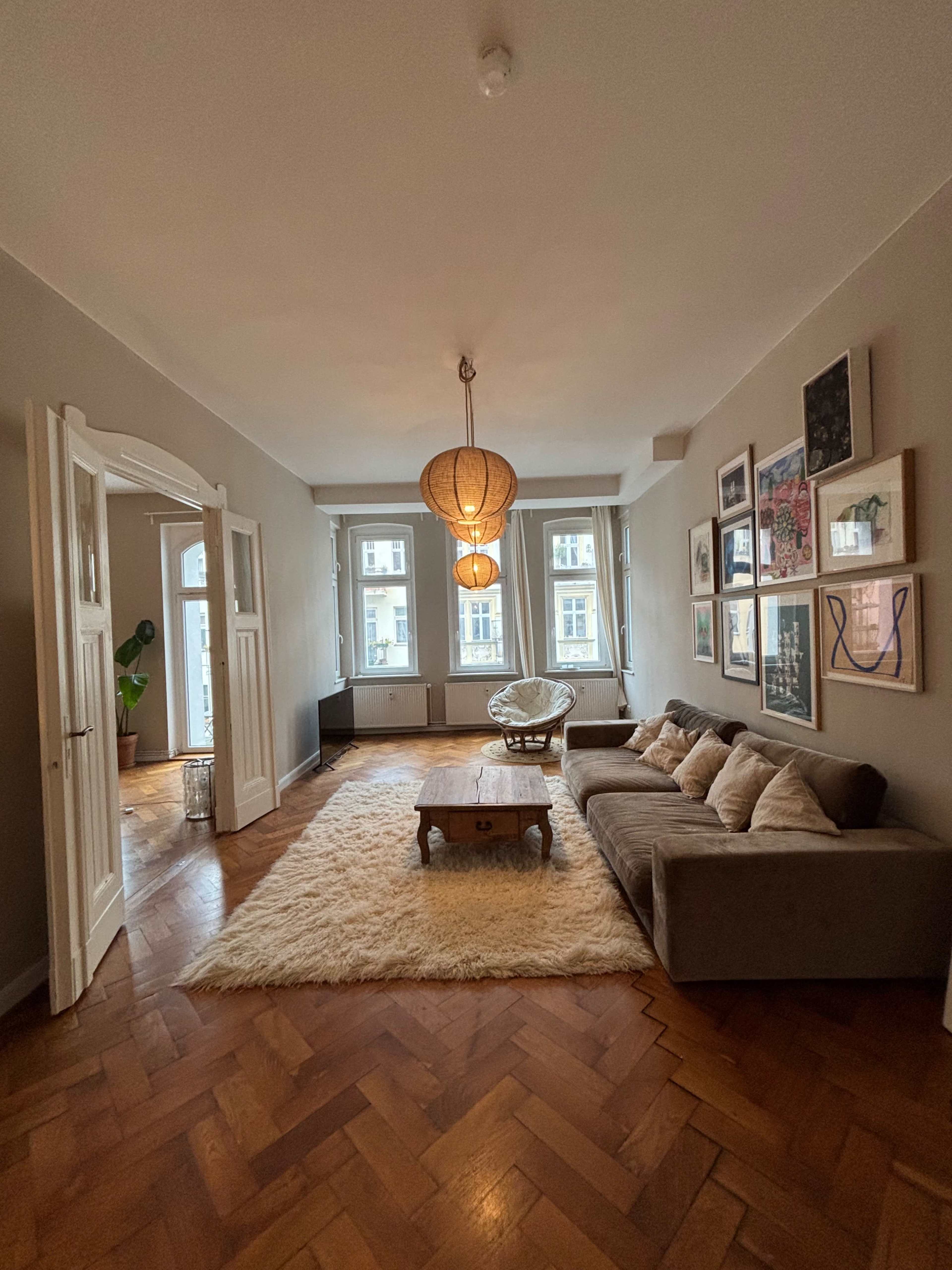 The image shows a spacious living room with a large rug, a sofa, a coffee table, and pendant lighting, with windows providing natural light.