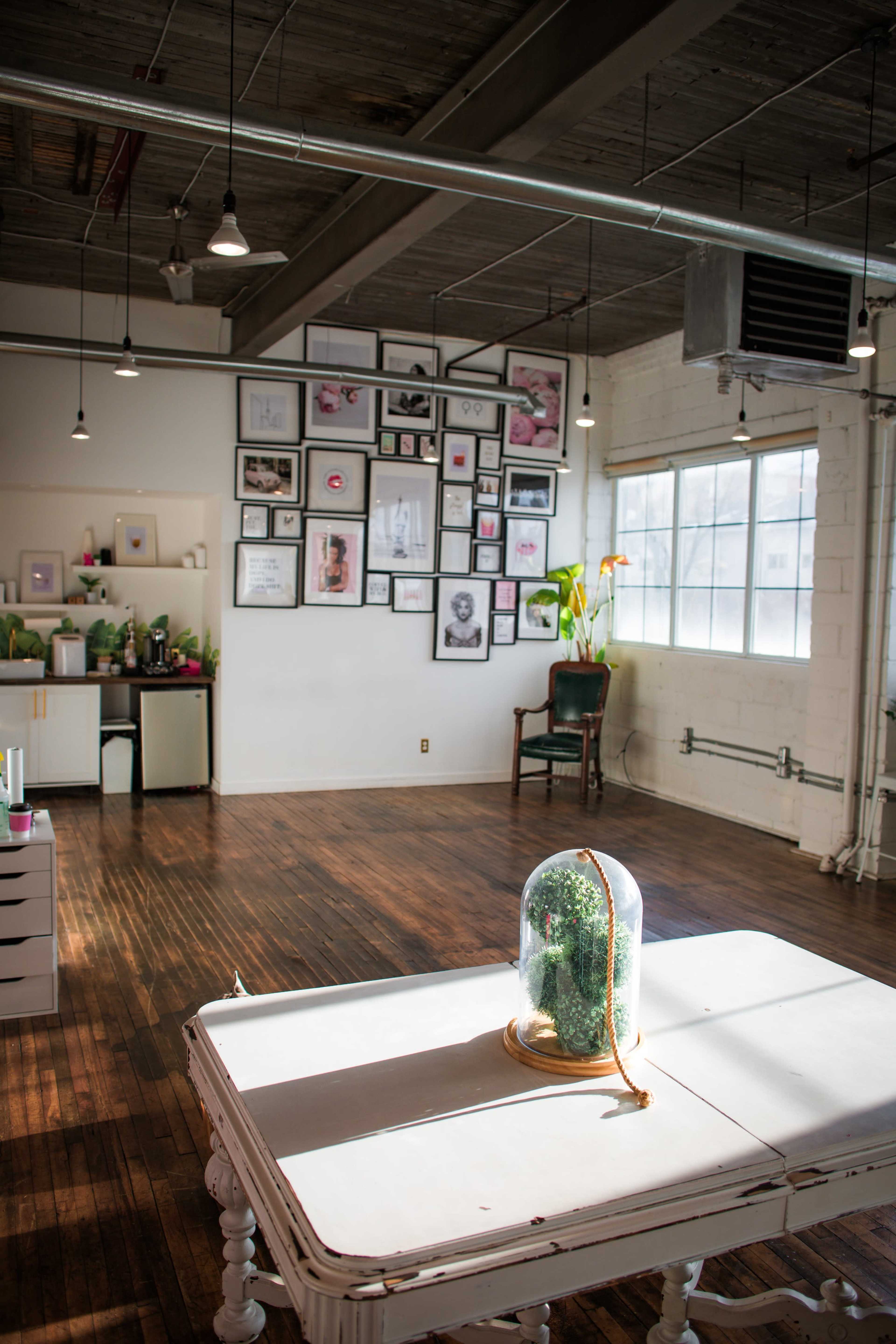 A bright, spacious room features a large window, hardwood floors, and a wall adorned with framed art, with a glass cloche holding greenery on a central table.