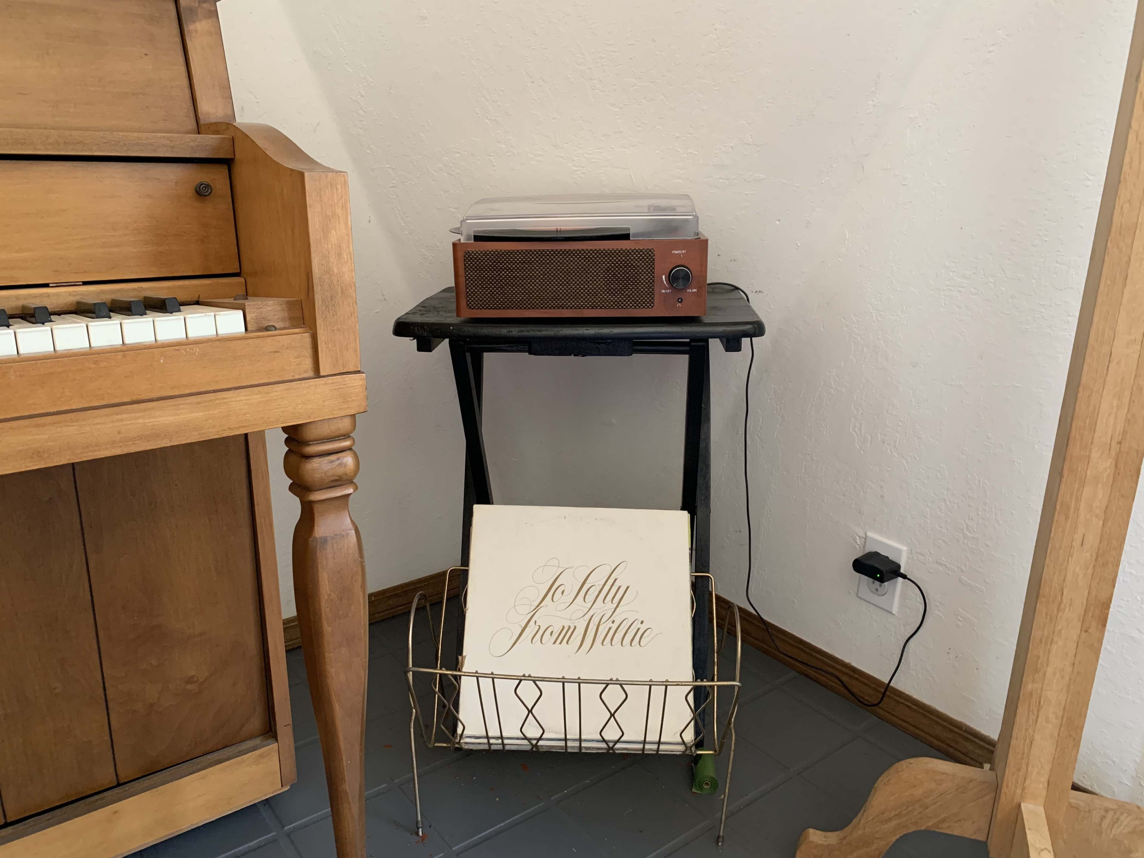 A wooden piano is positioned beside a record player on a small table, with a wire basket holding a record in the corner of the room.