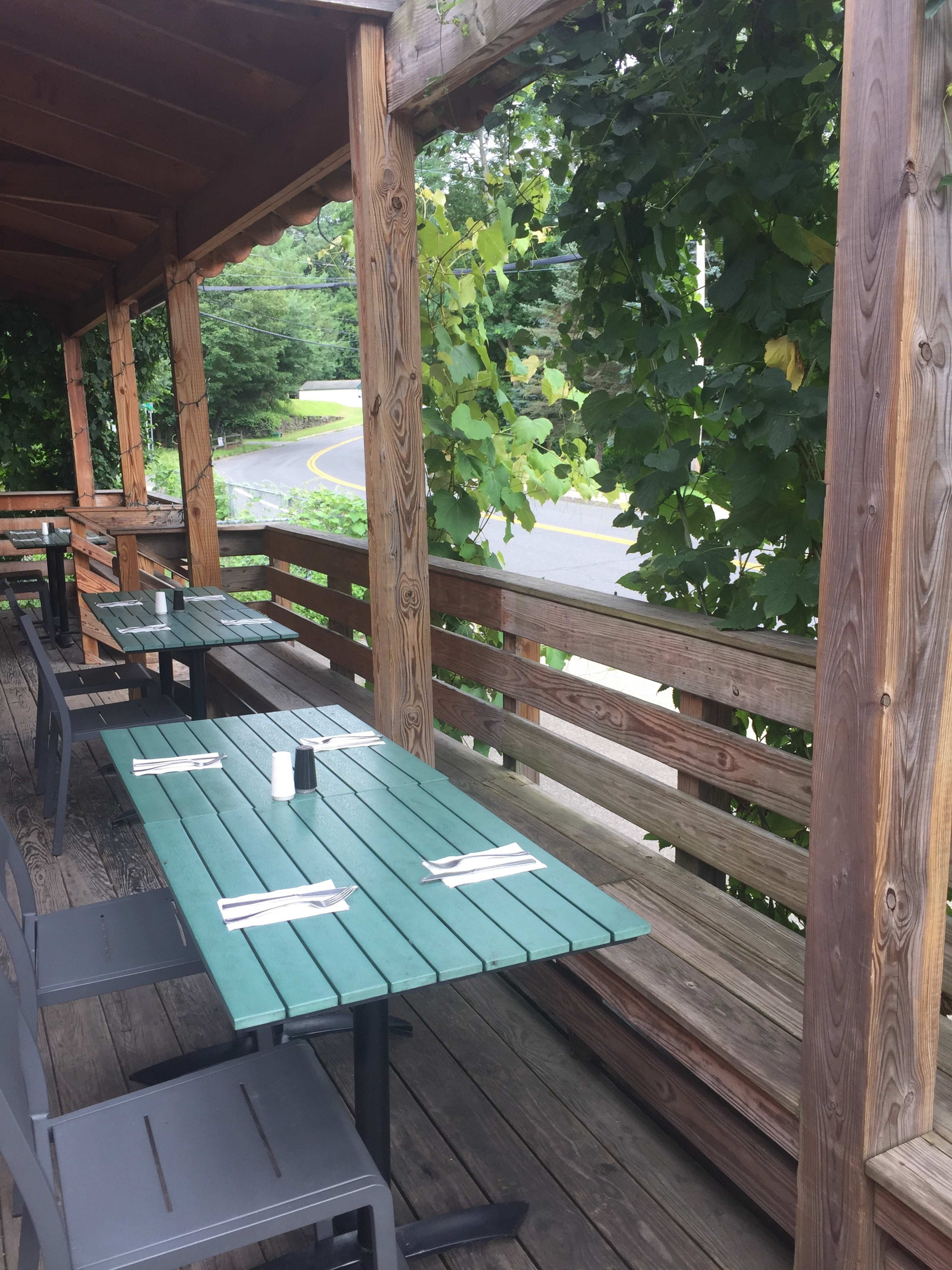 A patio area of a restaurant with wooden tables set for dining under a wooden awning, surrounded by greenery.