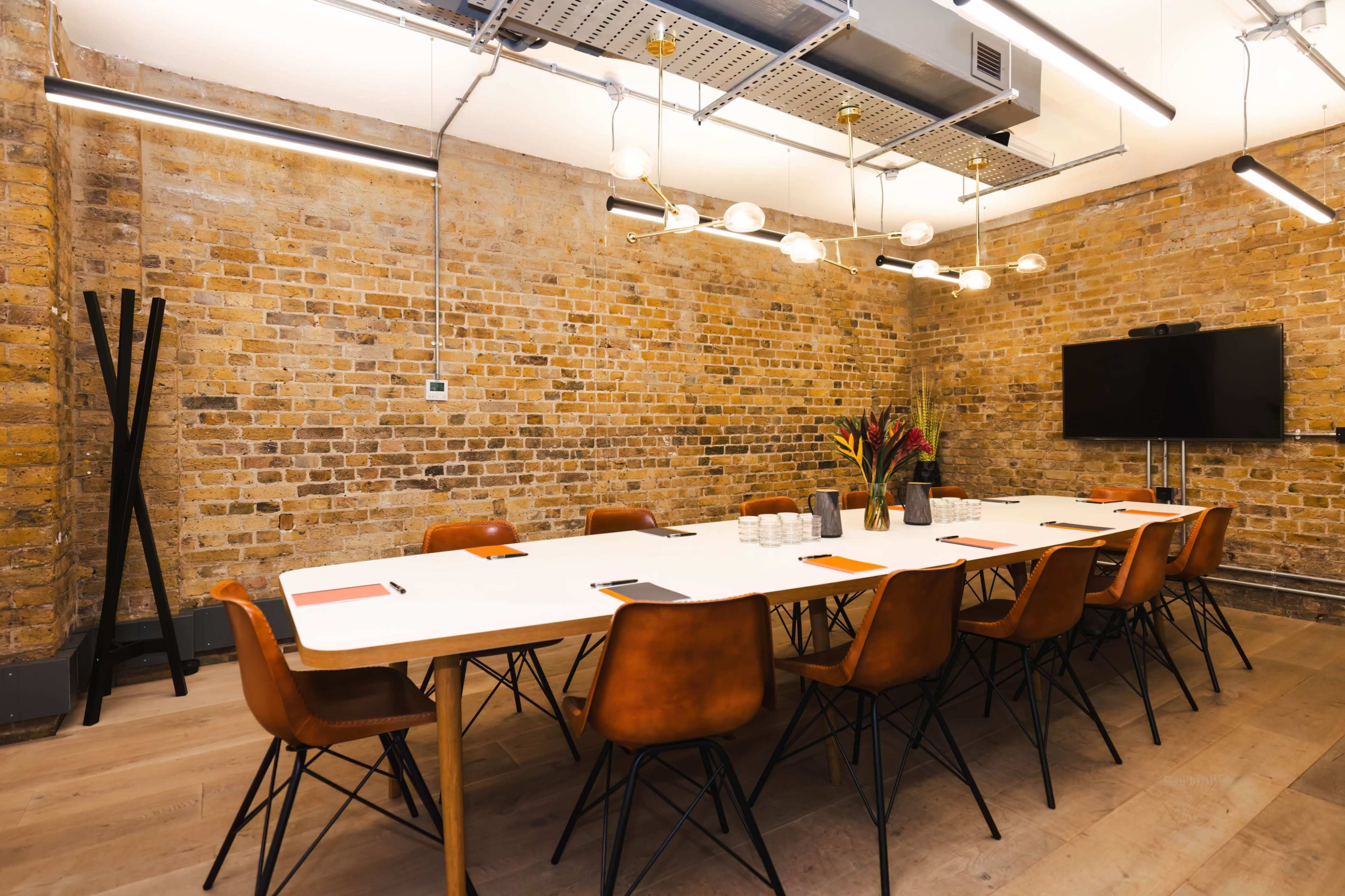 A modern conference room features a long white table surrounded by brown chairs, exposed brick walls, and overhead lighting.