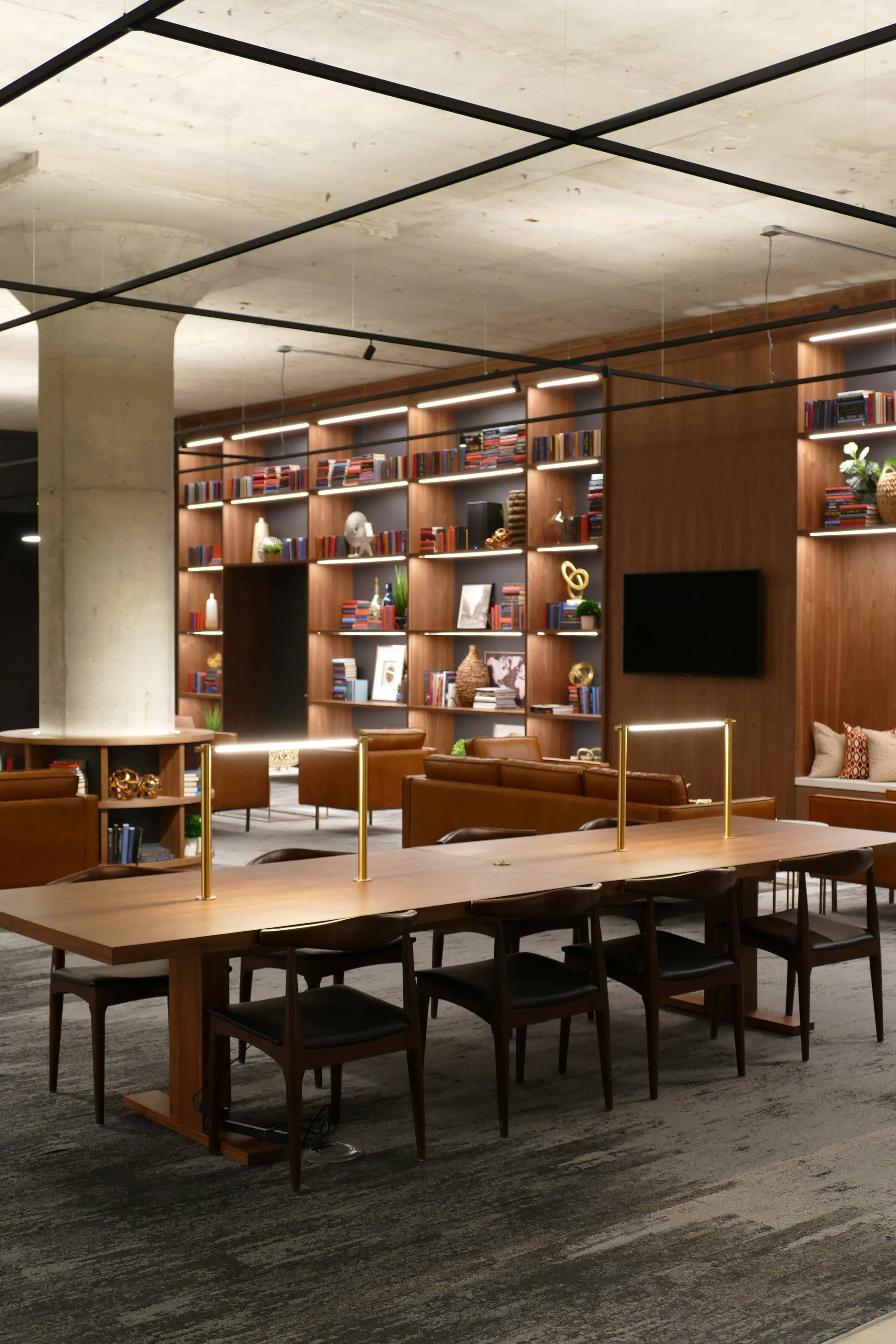 A modern office space features a long wooden table surrounded by chairs, with a backdrop of bookshelves filled with various items and a cozy seating area.