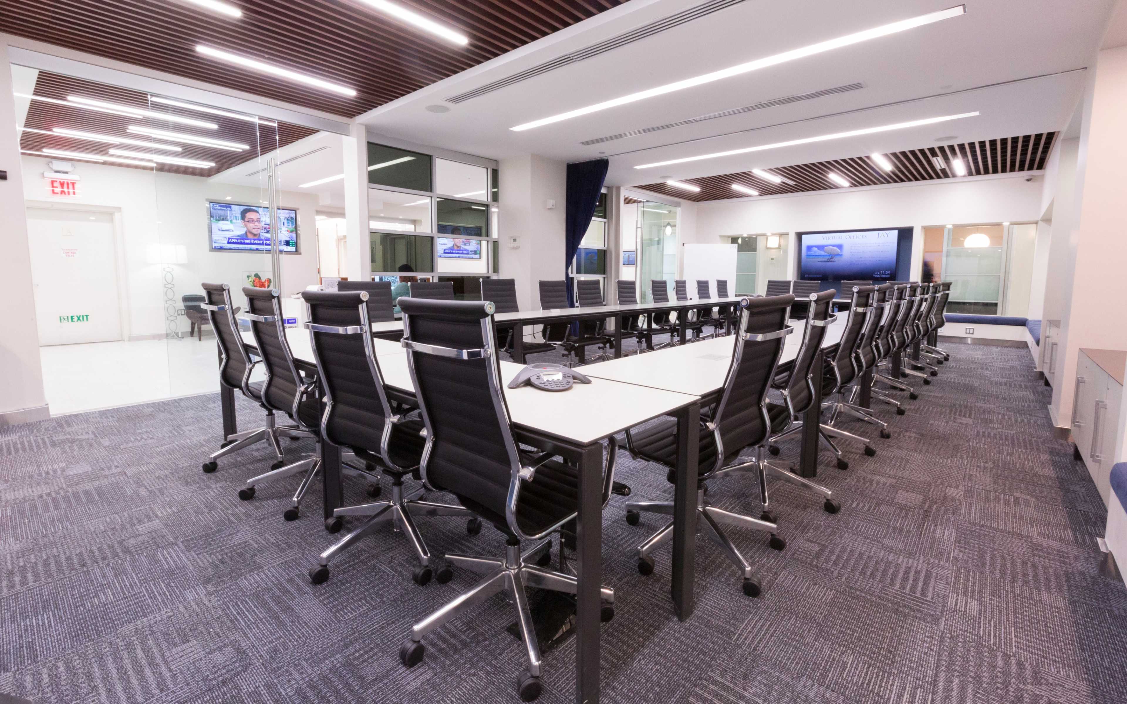 A long conference table surrounded by black office chairs is positioned in a well-lit meeting room featuring glass walls and multiple screens.