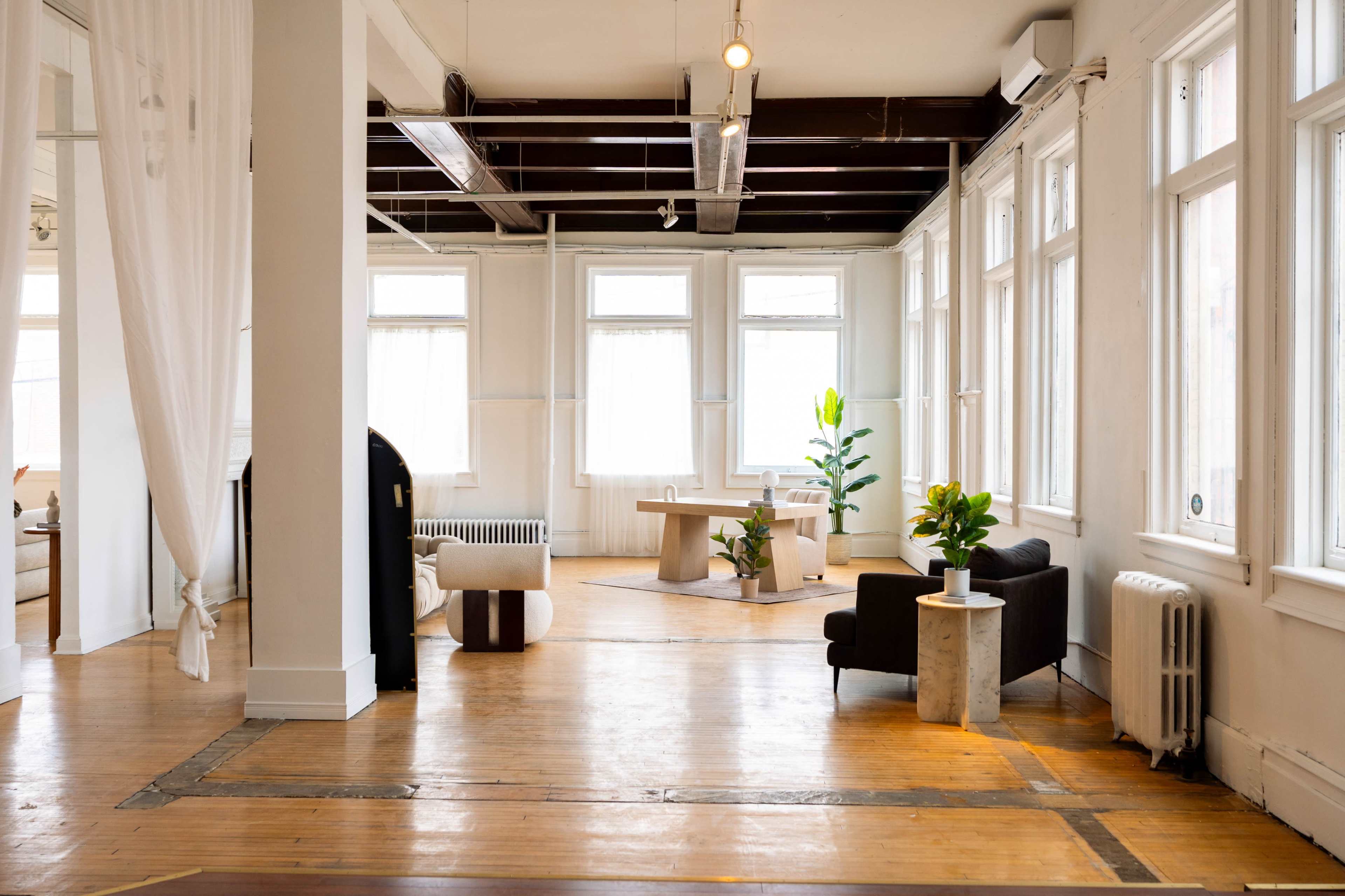 The image shows a spacious, well-lit room with large windows, featuring modern furniture, plants, and hardwood floors.