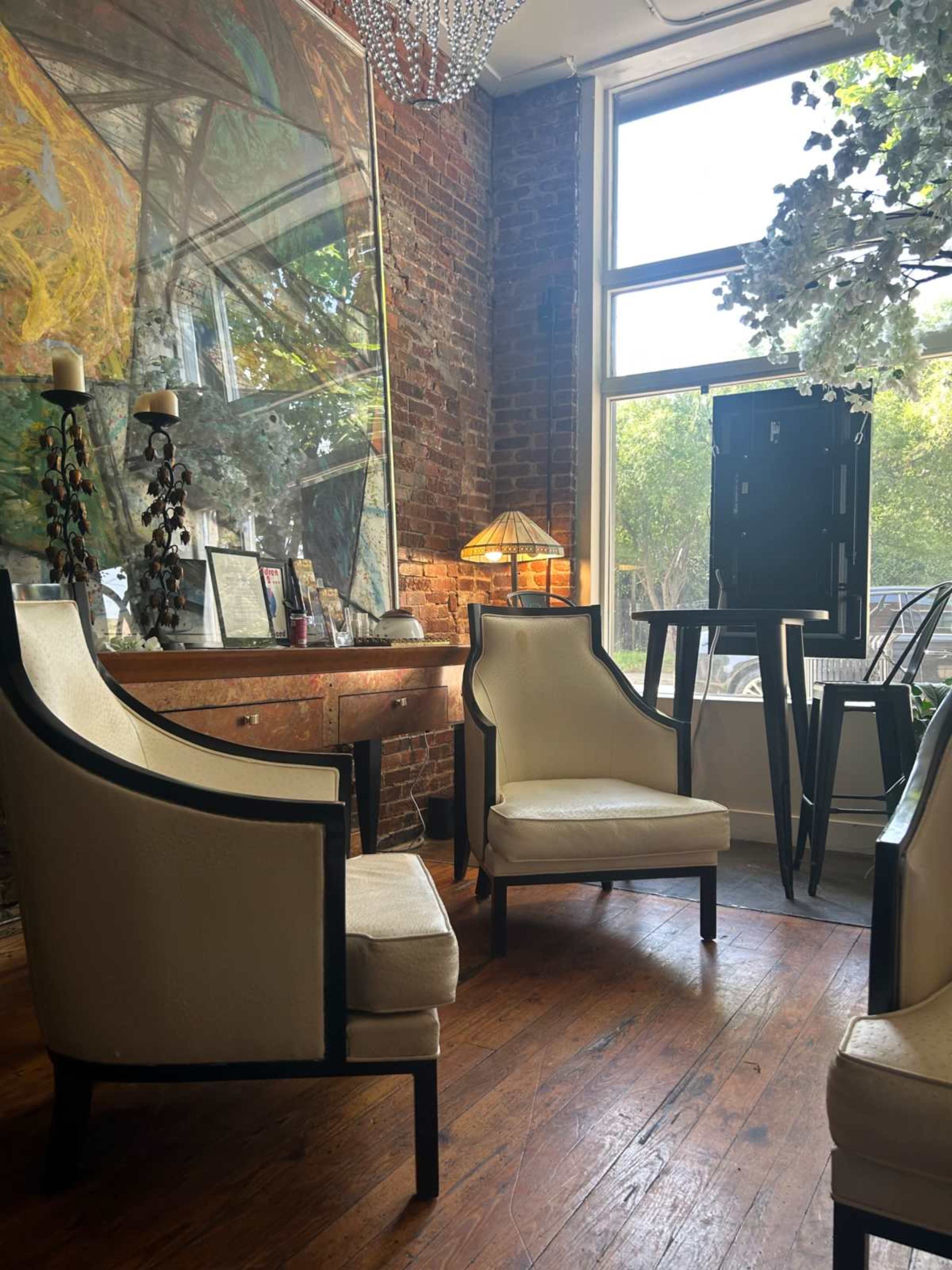 The scene shows a cozy corner of a room with two upholstered chairs, a small wooden table, and a large window allowing natural light to enter, along with a decorative painting on the brick wall.