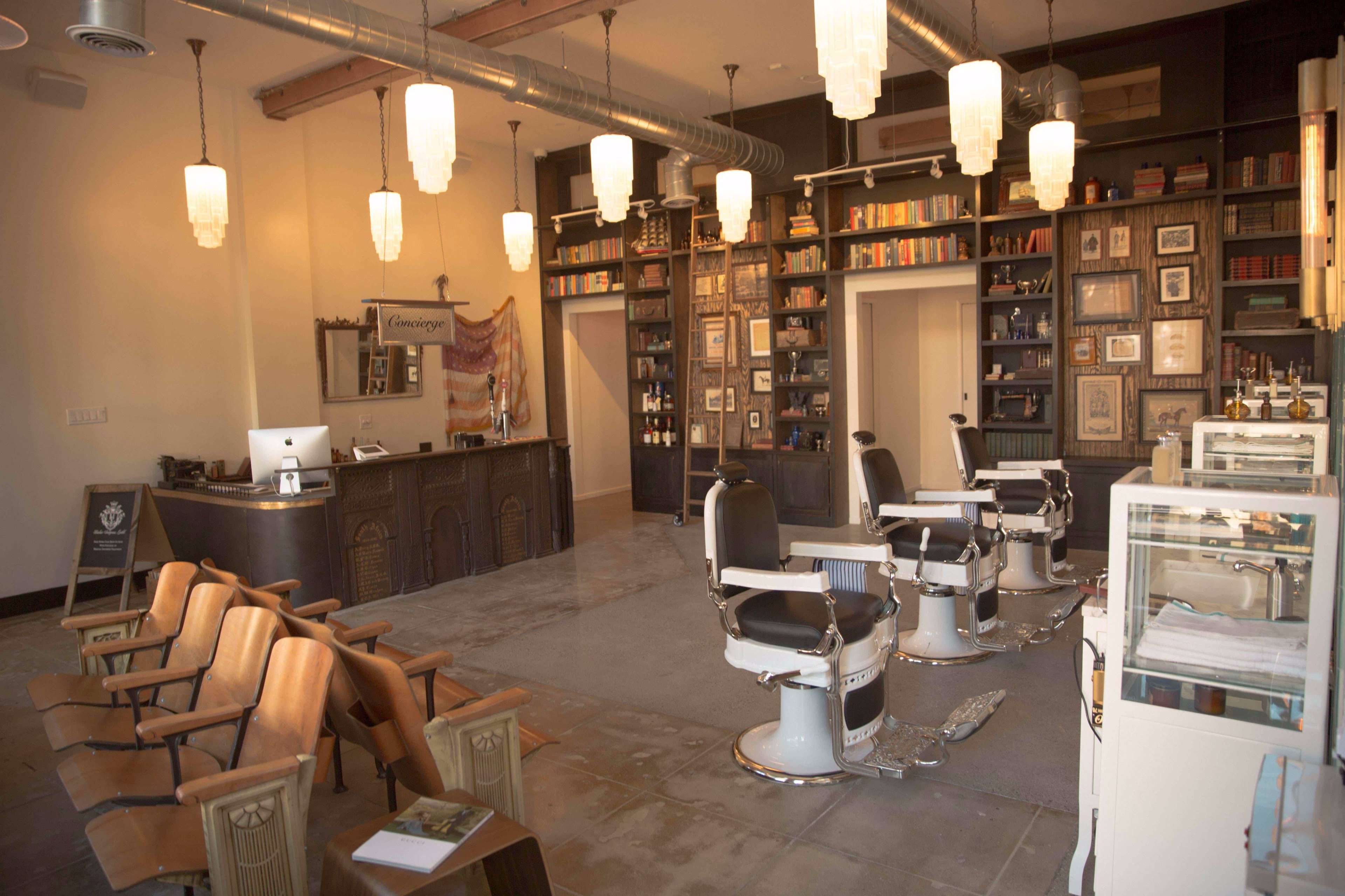 The interior of a stylish barber shop features several black barber chairs, a wooden reception desk with a computer, and shelves filled with books and decorative items.