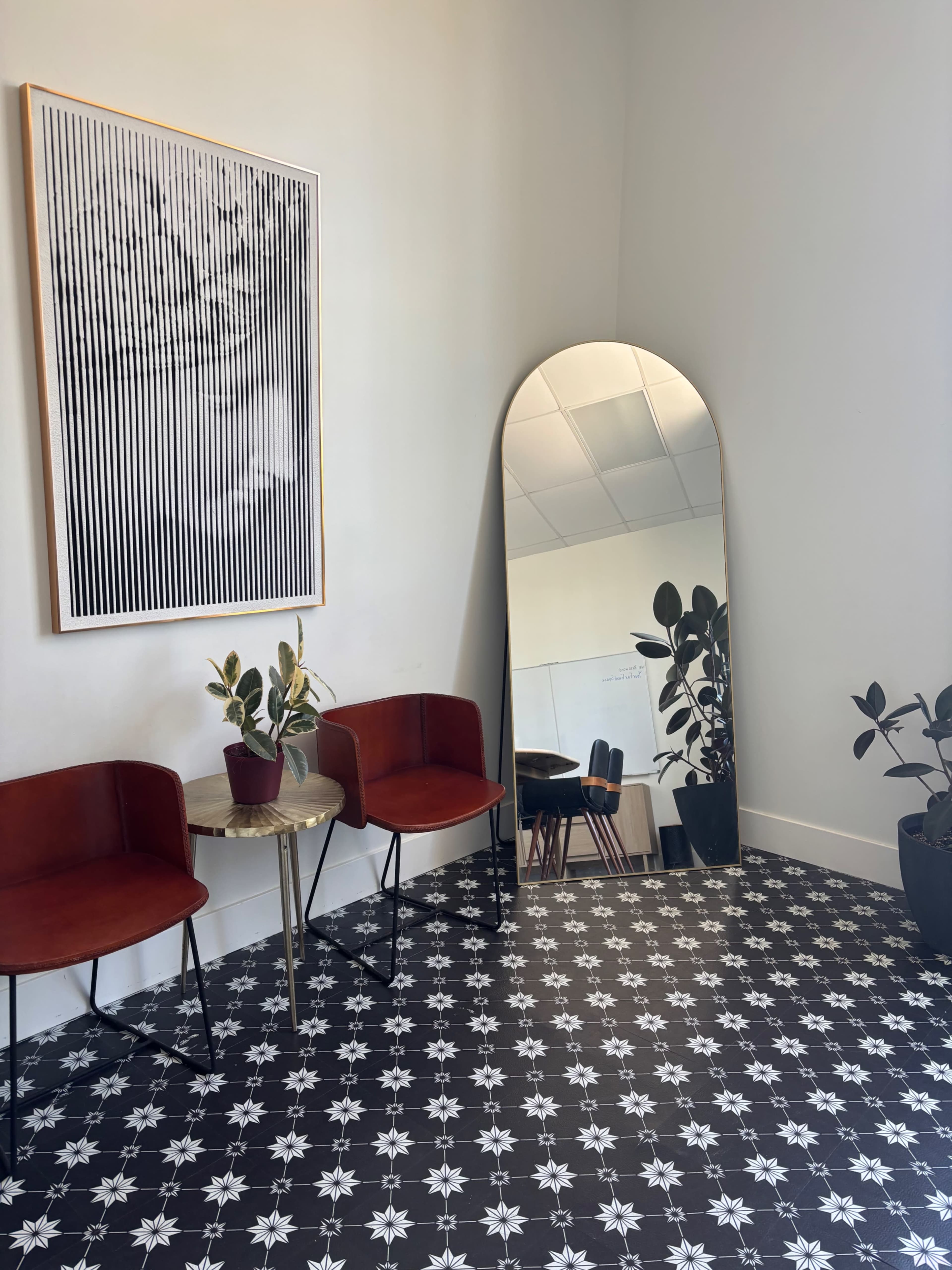 The image features a modern room with two red chairs, a round table with a plant, a large mirror, and patterned tile flooring.