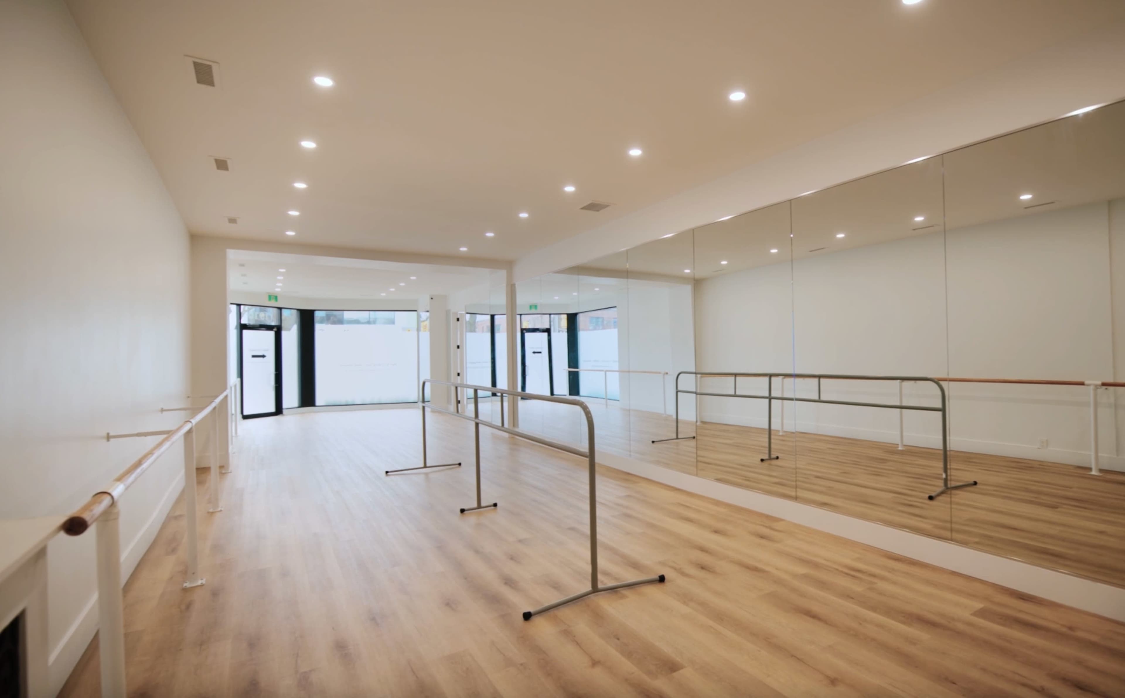 The image shows a light-filled ballet studio with wooden flooring, a mirror-lined wall, and a ballet barre along one side.