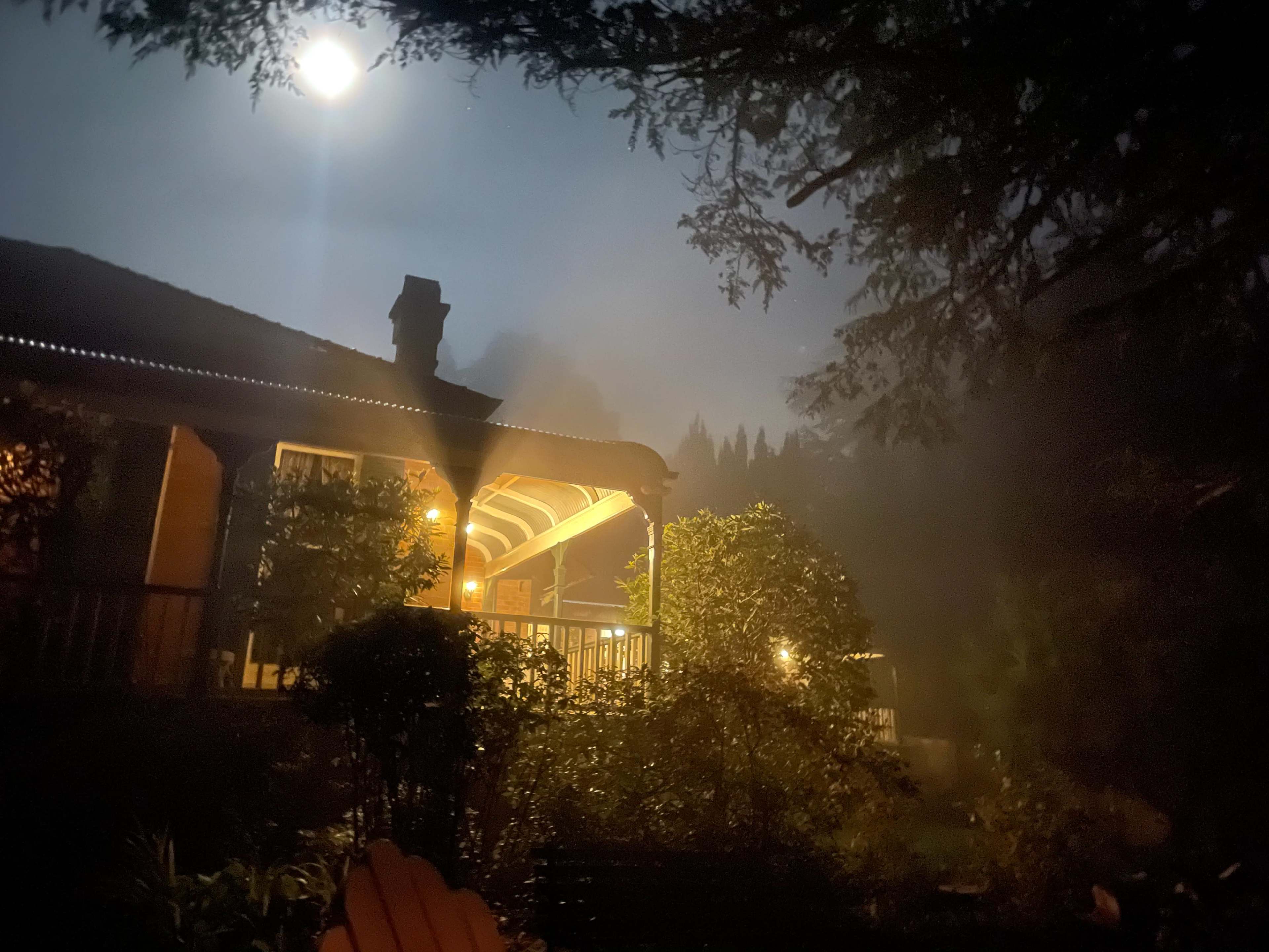 A well-lit porch stands in the foreground under a full moon, surrounded by fog and trees.