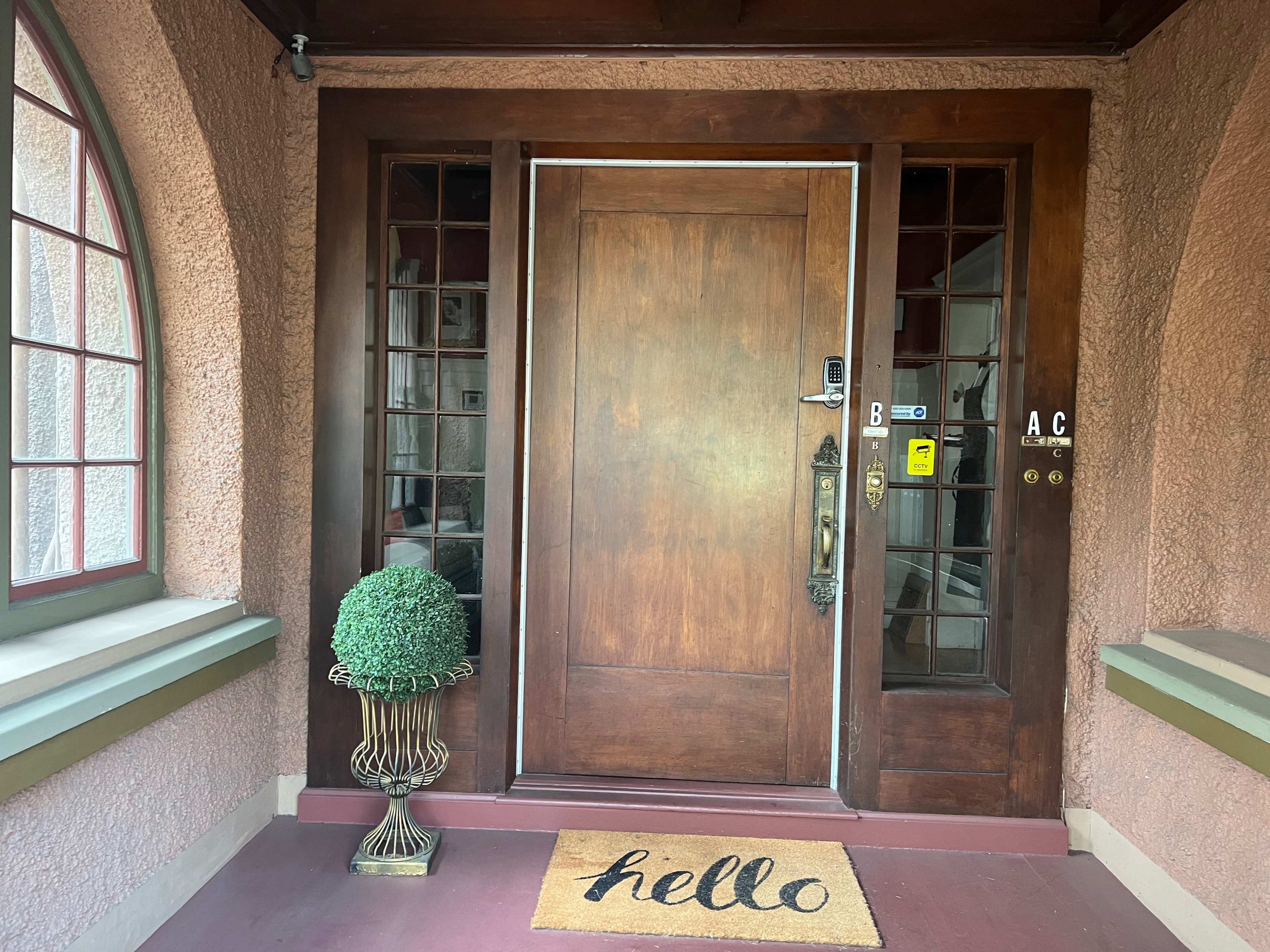 The image shows a wooden front door with glass panels on either side, a welcome mat, and a potted topiary beside it.