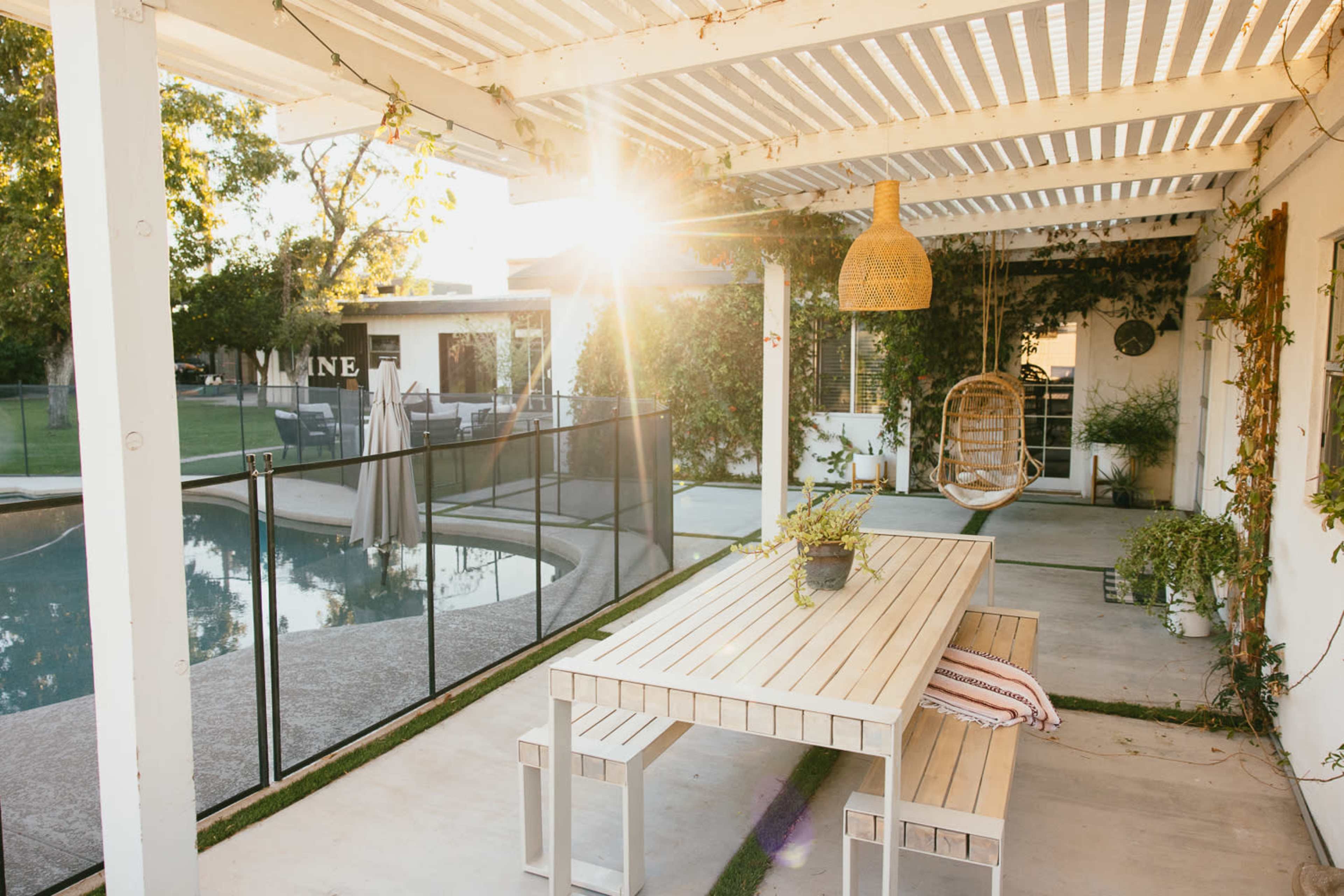 A modern outdoor patio features a wooden dining table near a swimming pool, with a hanging chair and lush greenery alongside.