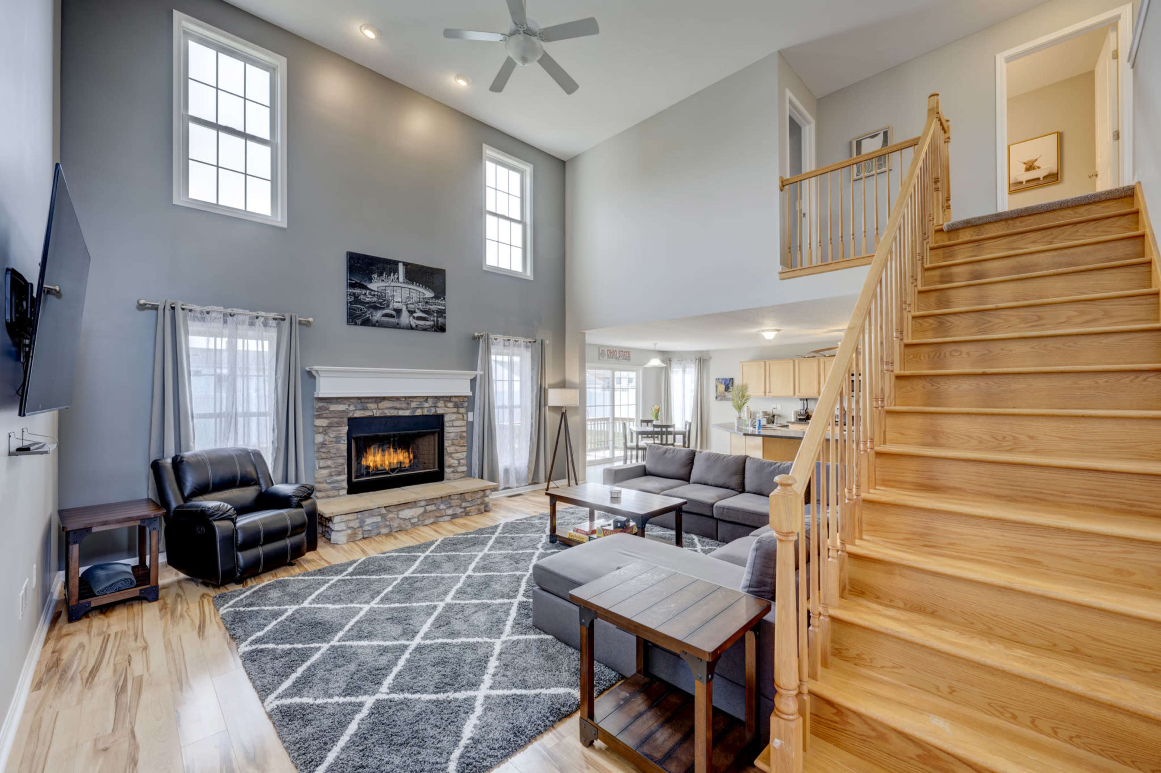 A two-story living room with a fireplace, large windows, a gray sectional sofa, and a wooden staircase leading to the upper level.