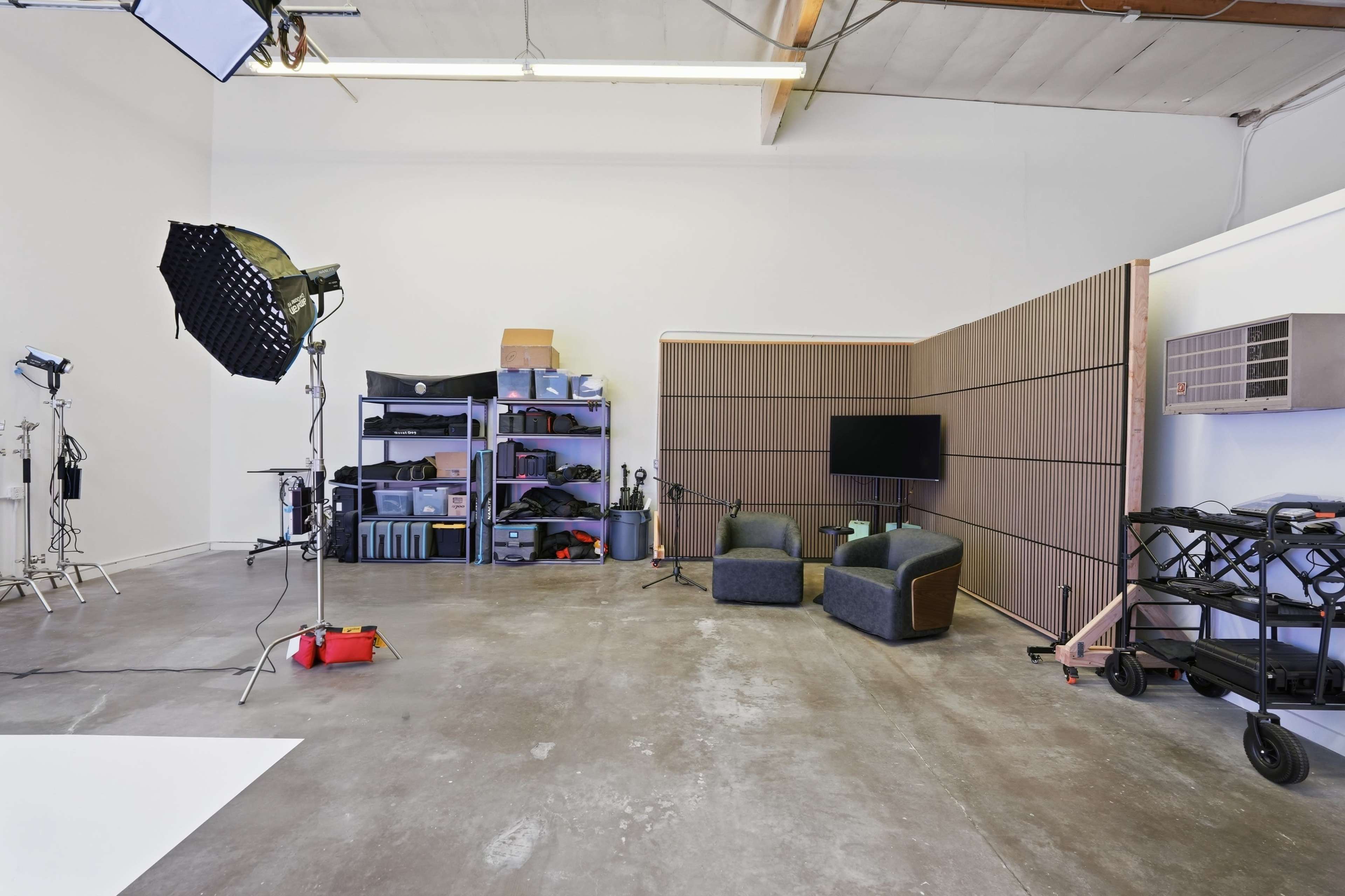 The image shows a spacious studio with a concrete floor, equipped with lighting equipment, storage shelves, a television setup, and two chairs.