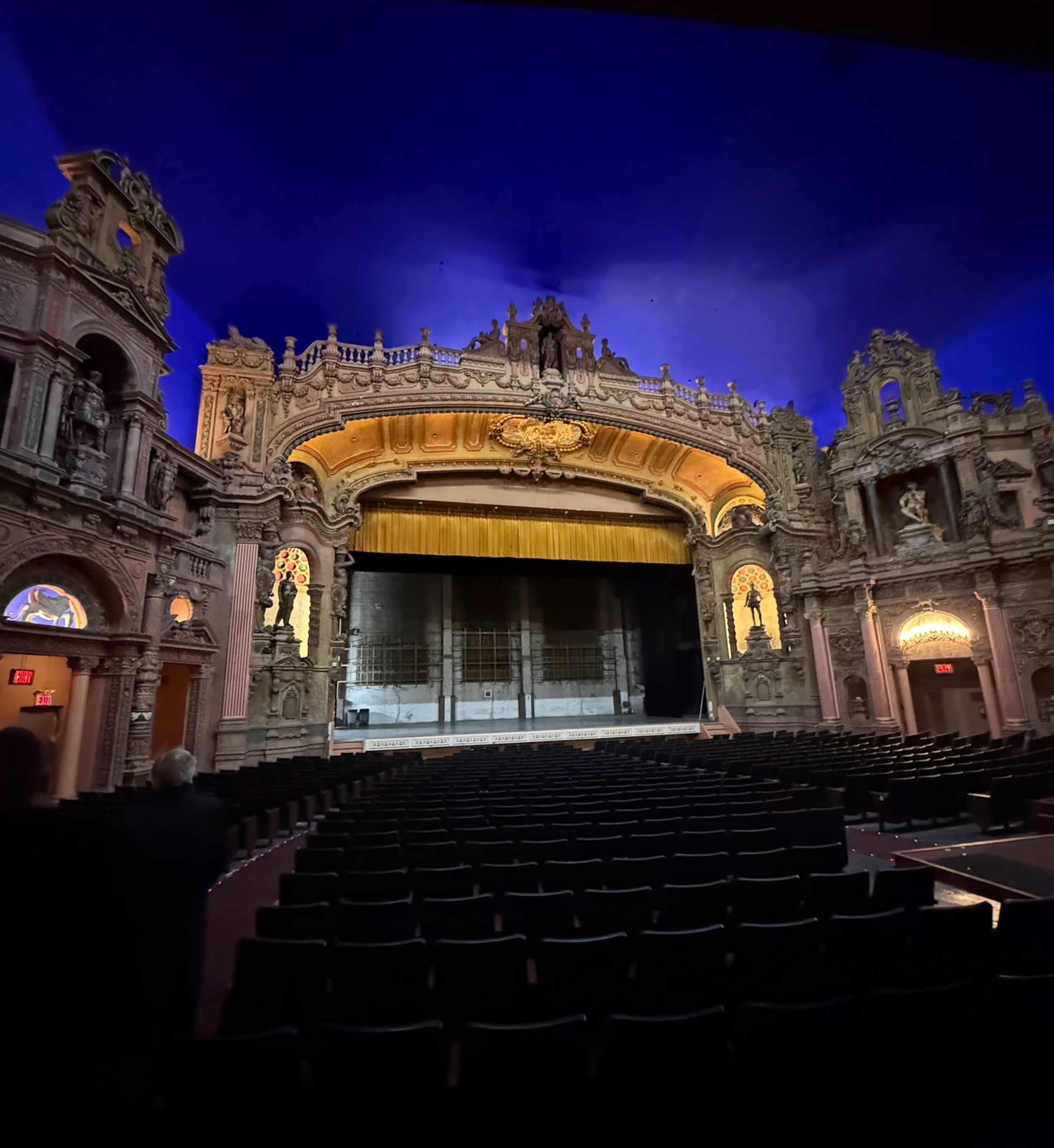 The image shows the interior of a grand theater with ornate architecture, featuring a stage with a golden curtain and rows of empty black seats.