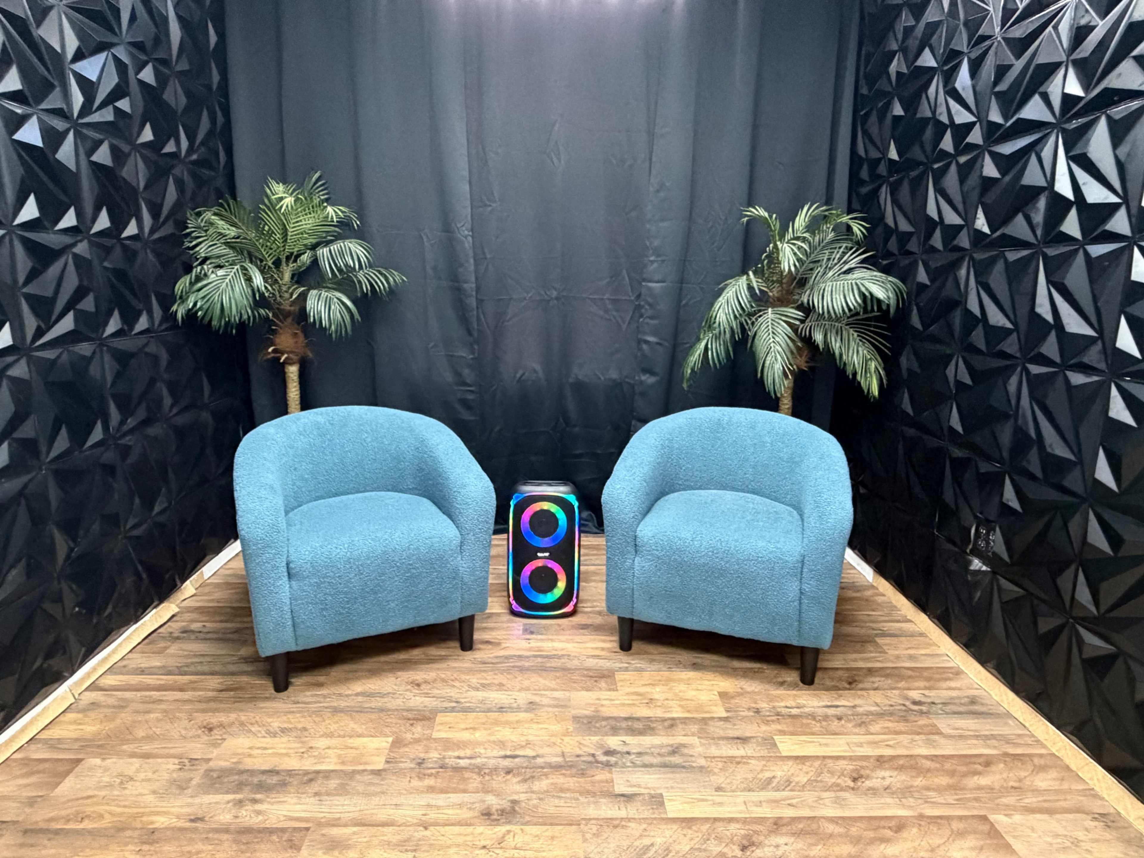 The image shows two blue chairs positioned opposite each other with two artificial palm trees and a colorful speaker in between, against a dark, textured backdrop.