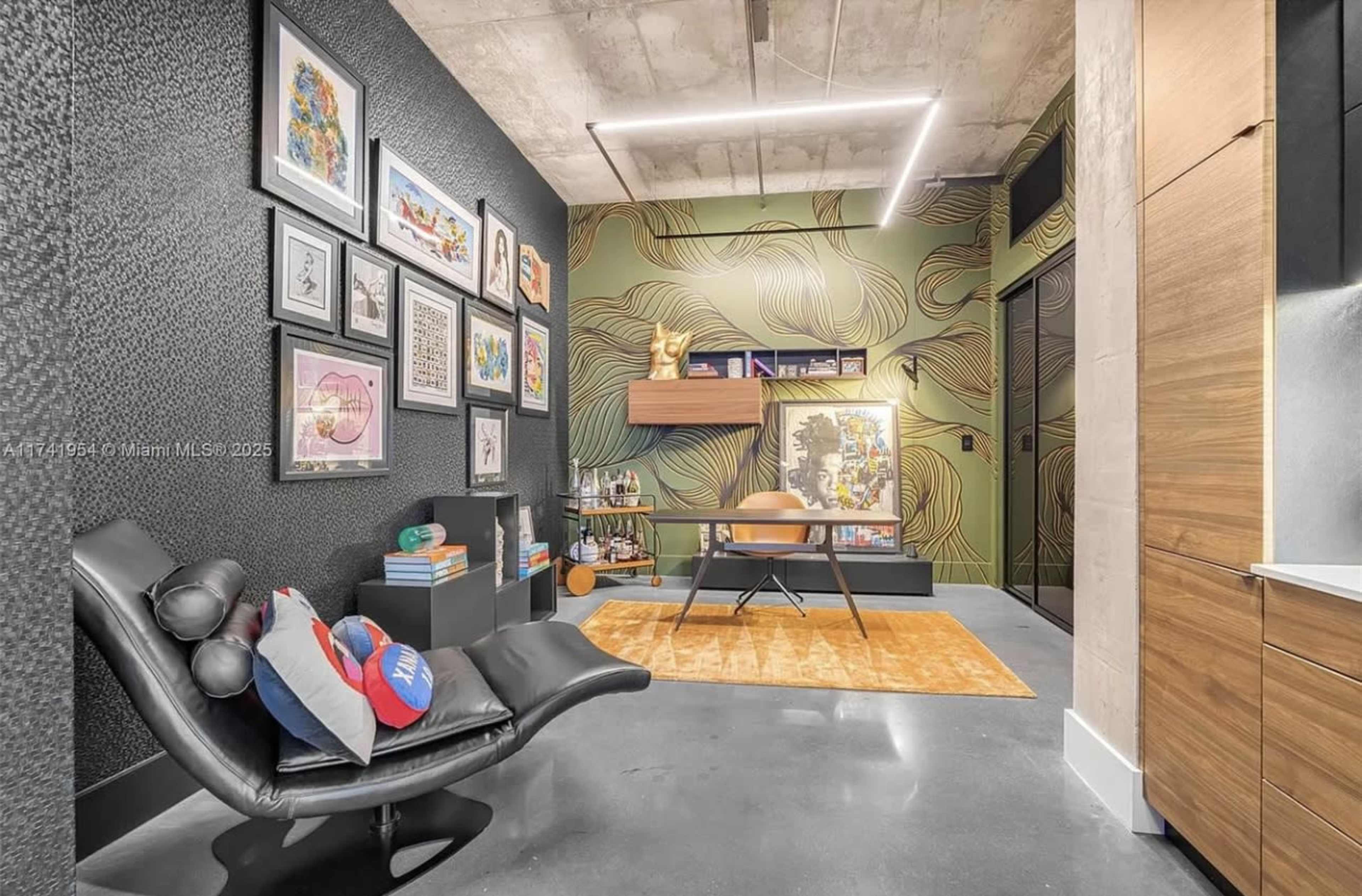 A modern home office features a black accent wall with framed art, a wooden desk, a lounge chair, and a decorative rug on a polished concrete floor.