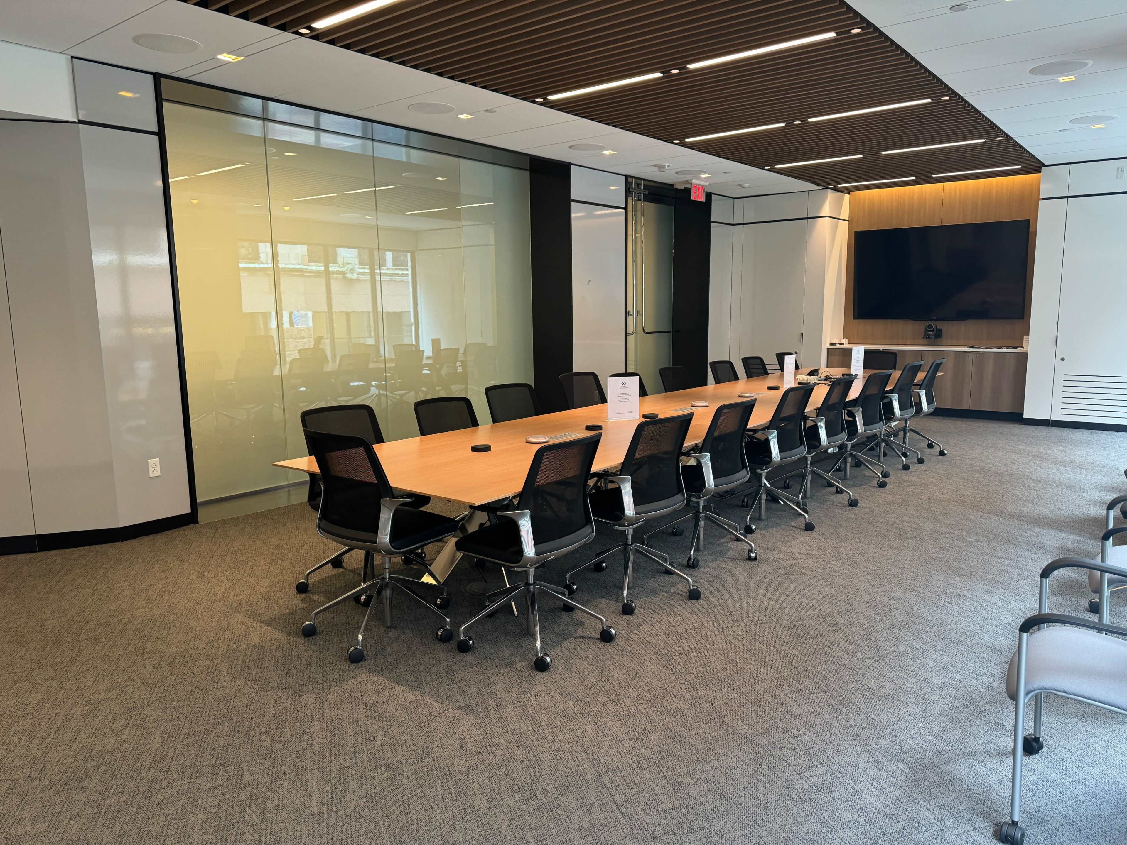A spacious conference room features a long wooden table surrounded by black office chairs and a wall-mounted screen in the background.