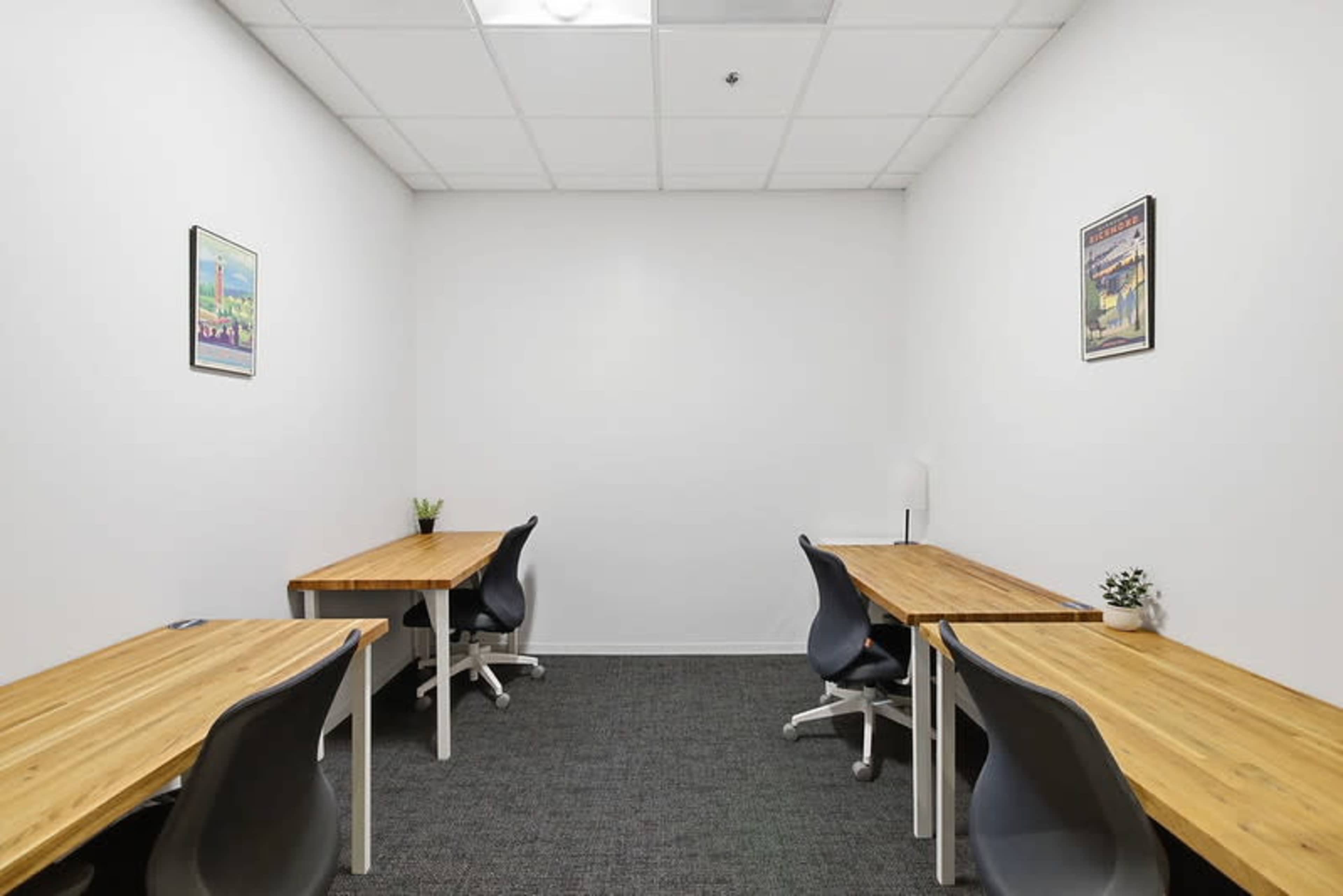 The image shows a small, minimalistic office space with two wooden desks and chairs, separated by a neutral-colored wall.