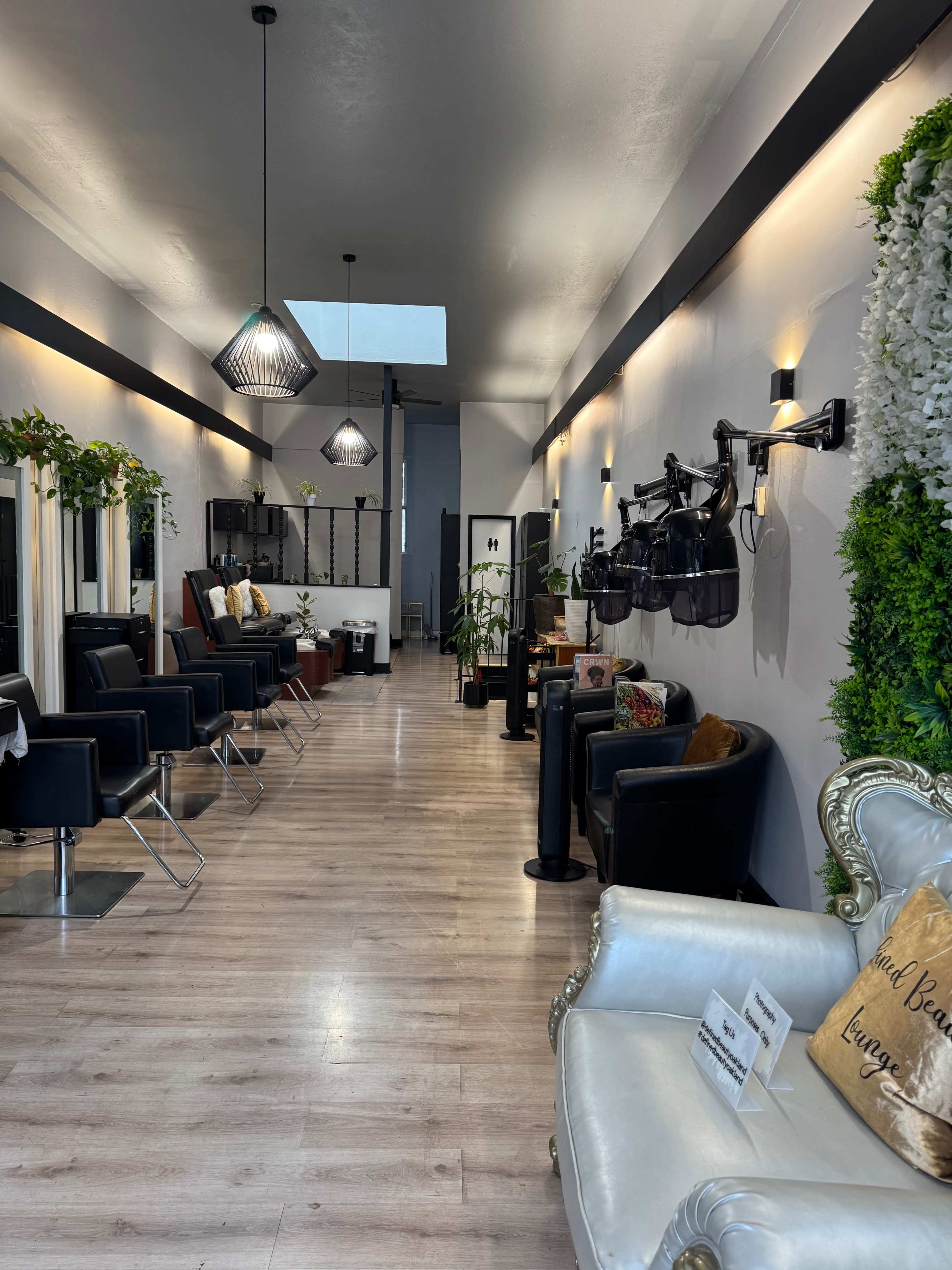The image shows a well-lit salon interior featuring several styling stations, comfortable seating, and decorative plants along the walls.