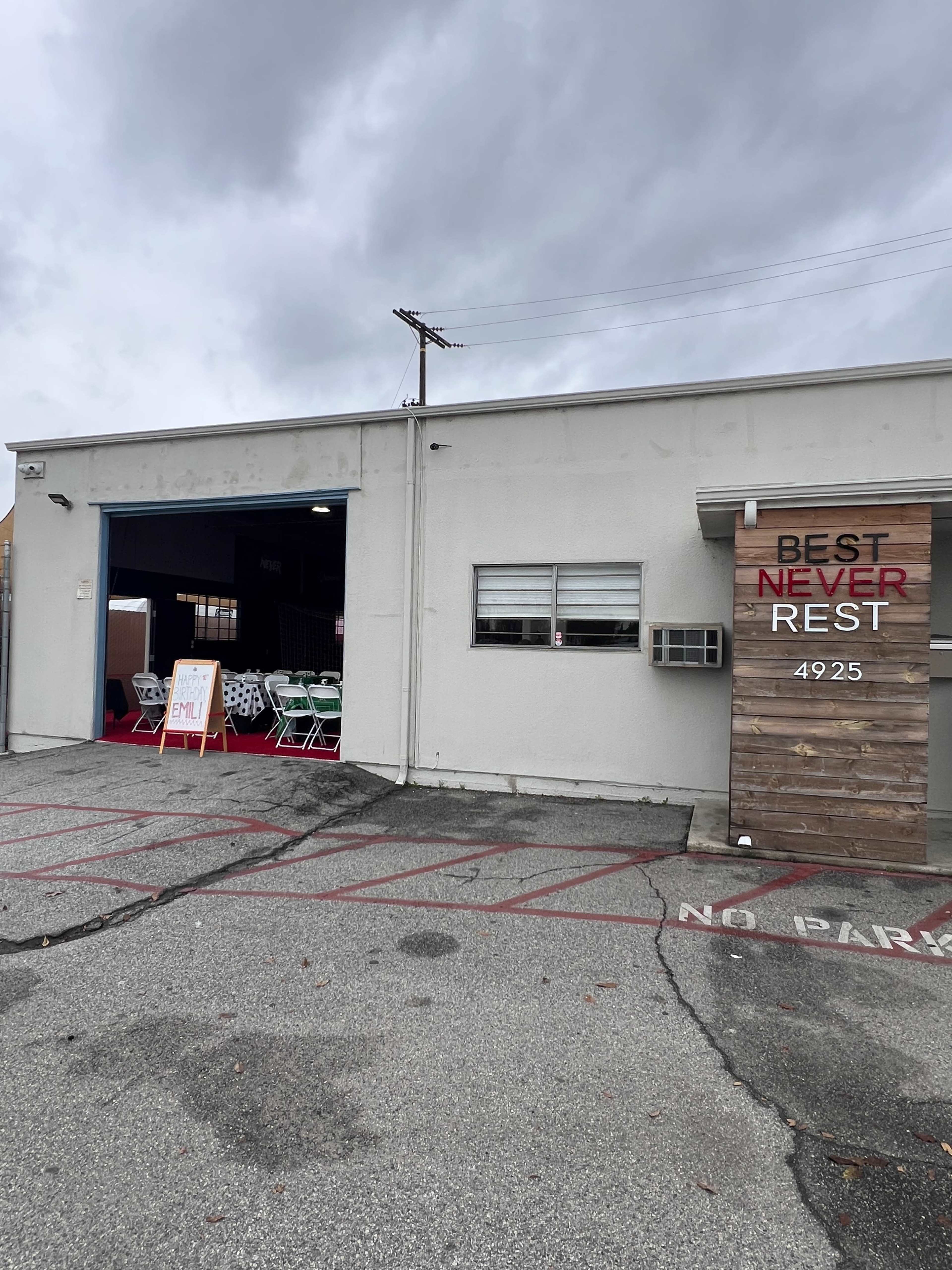 The image shows a building with a gray exterior, featuring an open garage door and a wooden sign that reads "BEST NEVER REST" beside the entrance.