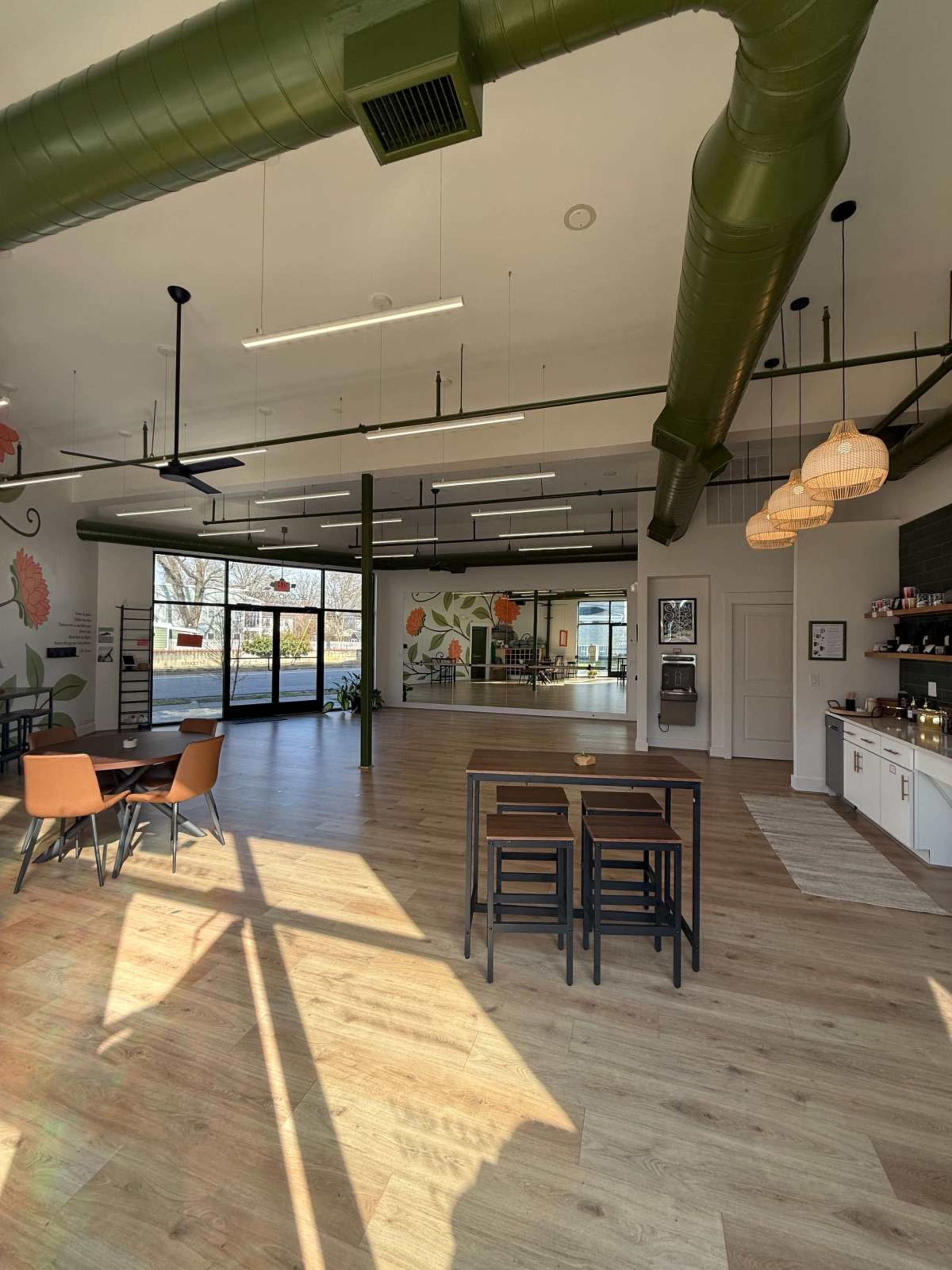 The image shows a spacious, well-lit interior of a café or community space with wooden floors, green ceiling ducts, and large windows letting in natural light.