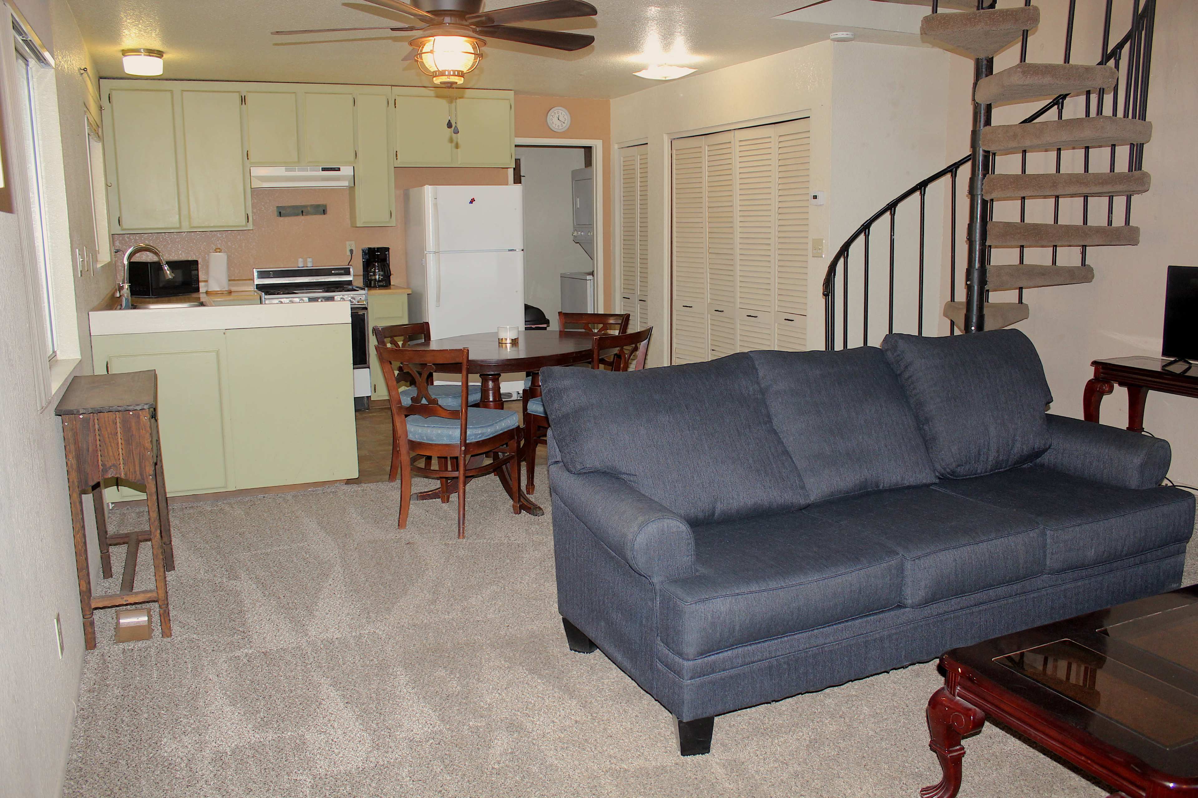 A living area with a blue couch, a dining table with chairs, a kitchen in the background, and a spiral staircase.