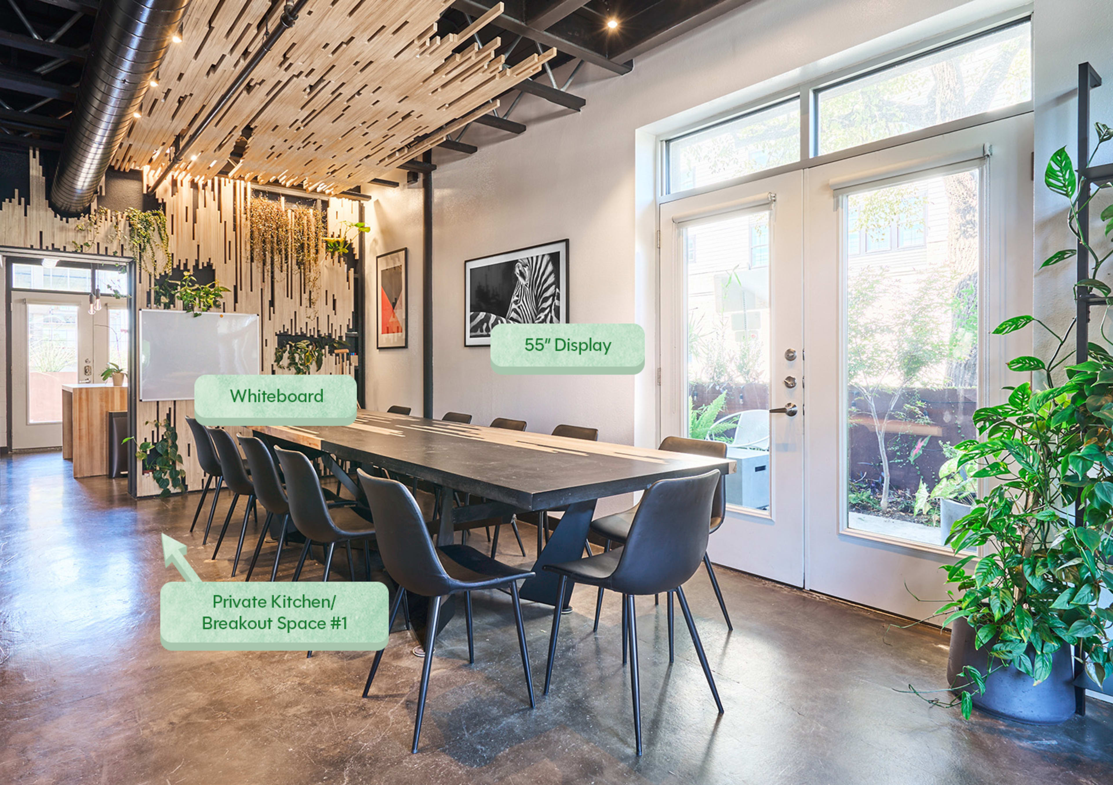 A modern meeting room features a long black table surrounded by chairs, a whiteboard, a 55-inch display, and large windows leading to an outdoor area, adorned with greenery.