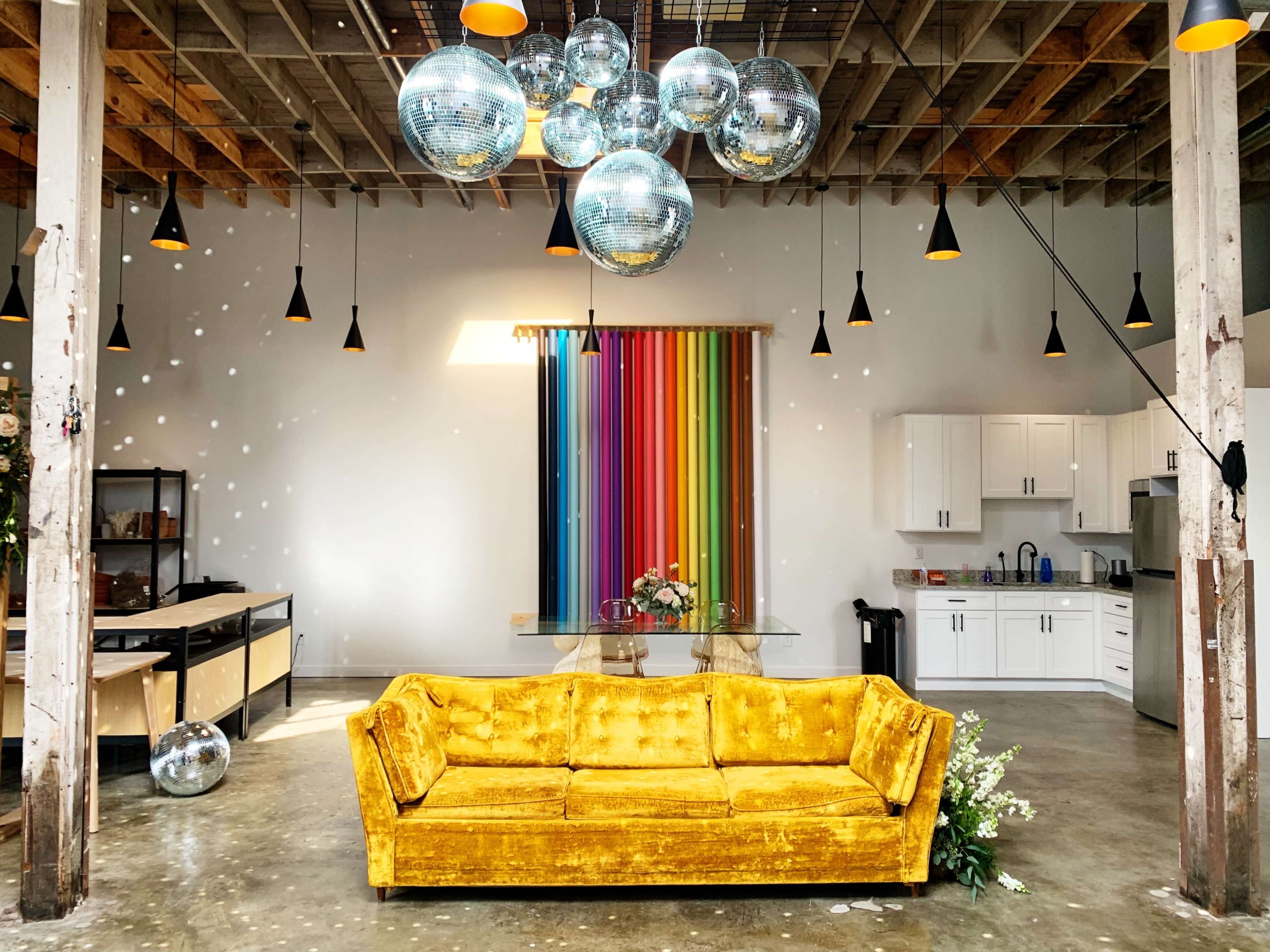 A yellow velvet couch is centered in a spacious room with a colorful striped backdrop, disco balls hanging from the ceiling, and a modern kitchen in the background.