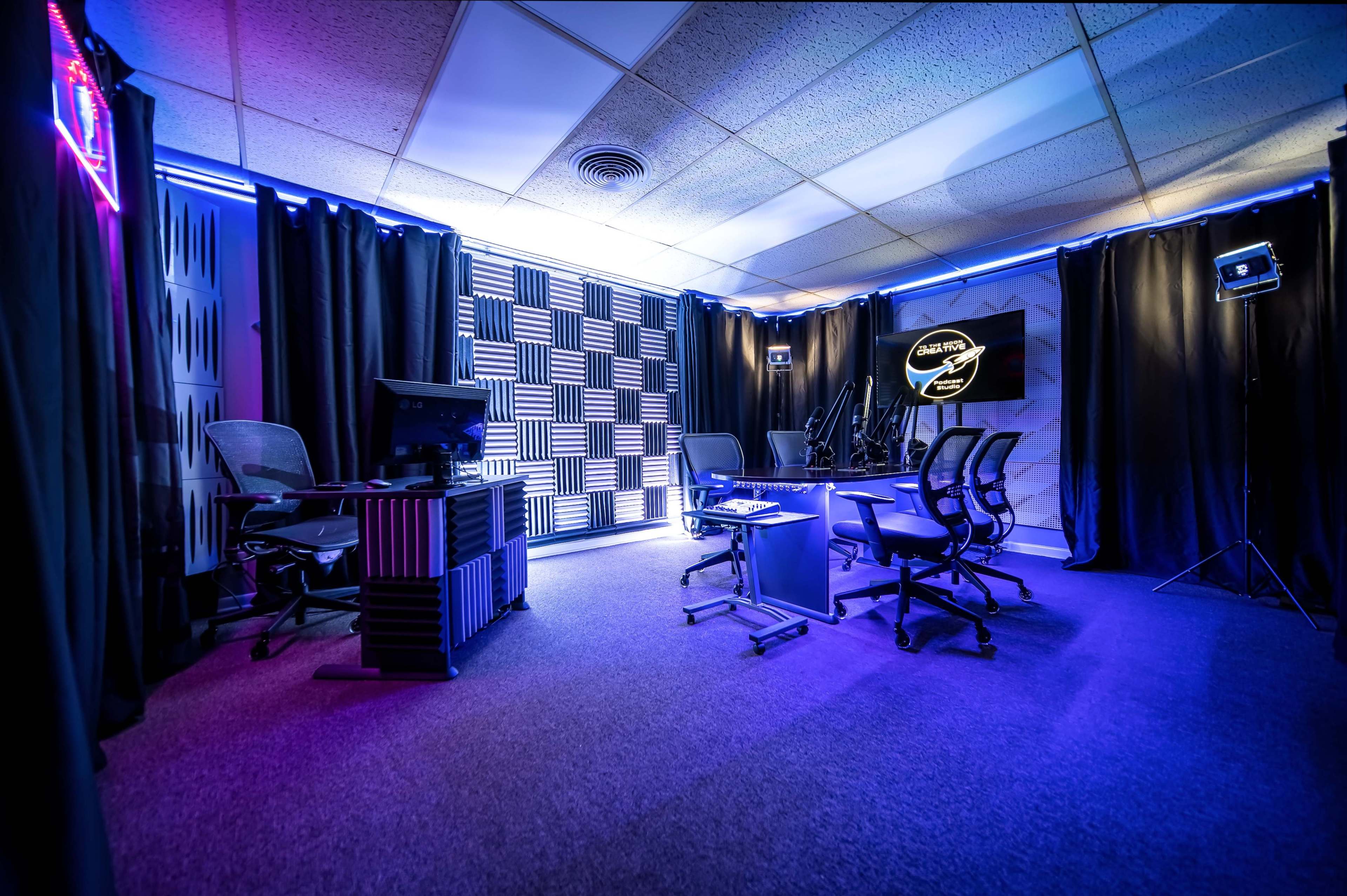 A modern studio featuring black curtains, a checkered wall, and four chairs arranged around a central table under blue and purple lighting.