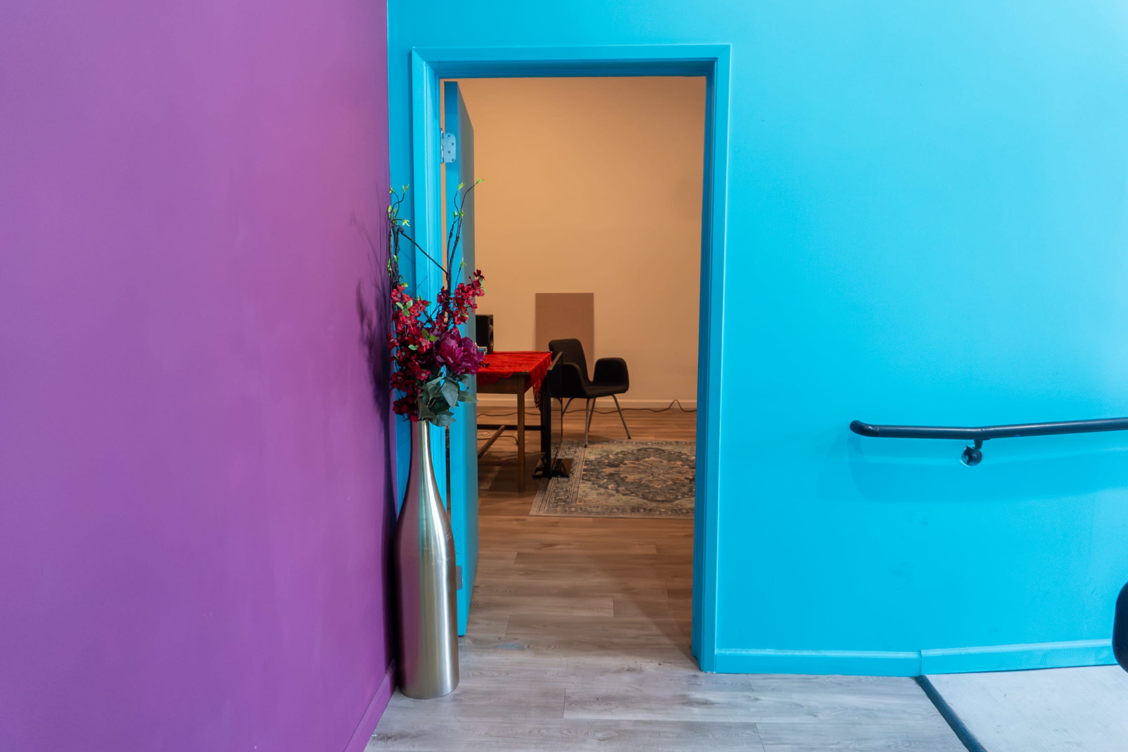 A doorway framed by purple and blue walls opens into a room with a table and a chair in the background.